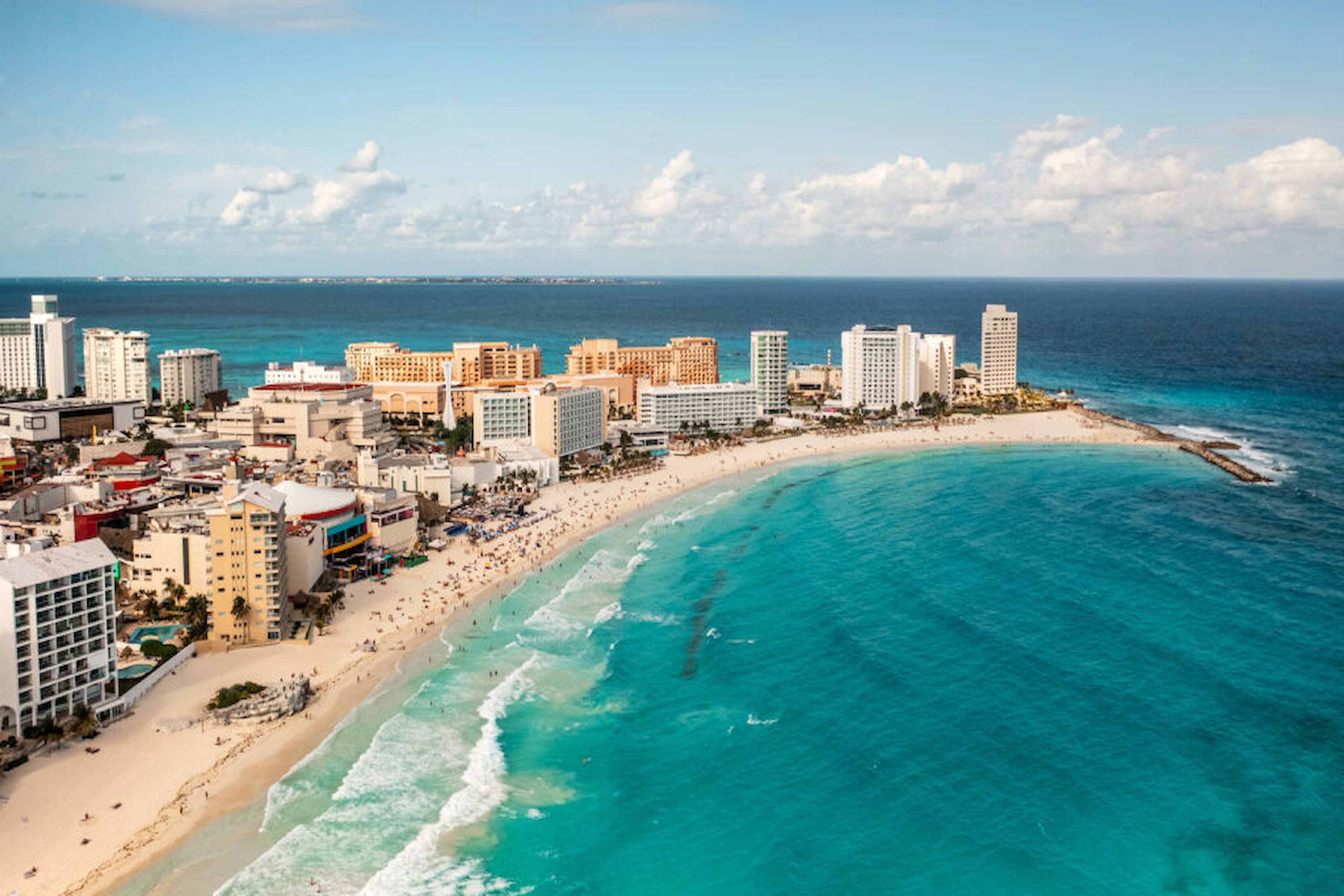 Aerial view of a coastal city with high-rise buildings along a curved sandy beach, bordered by clear turquoise water under a blue sky with scattered clouds.