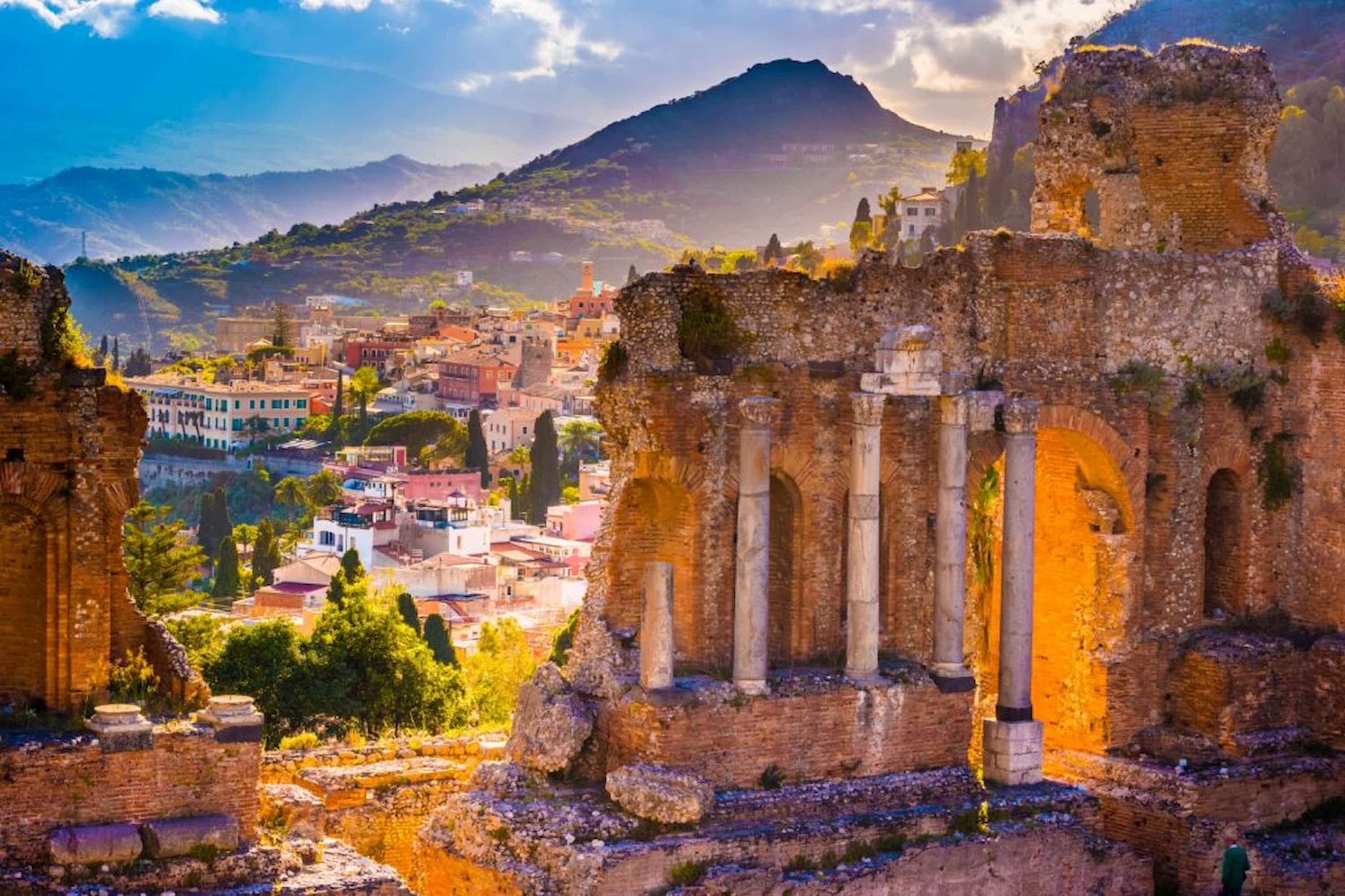 Ancient stone ruins with arches, overlooking a vibrant town and mountainous landscape under a cloudy sky, illuminated by golden sunlight.