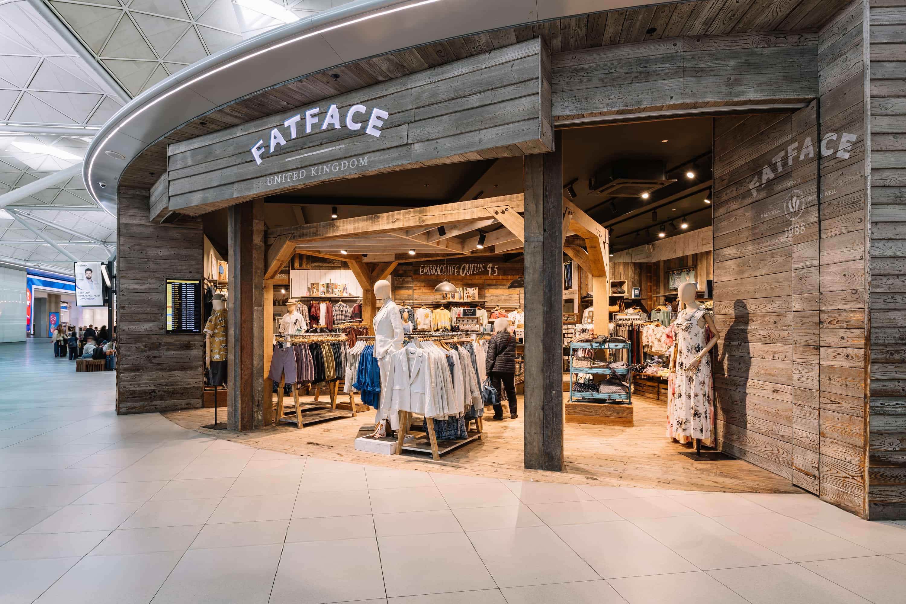 Wooden storefront of a clothing store with mannequins and racks of clothes inside, located in a spacious, modern airport terminal.