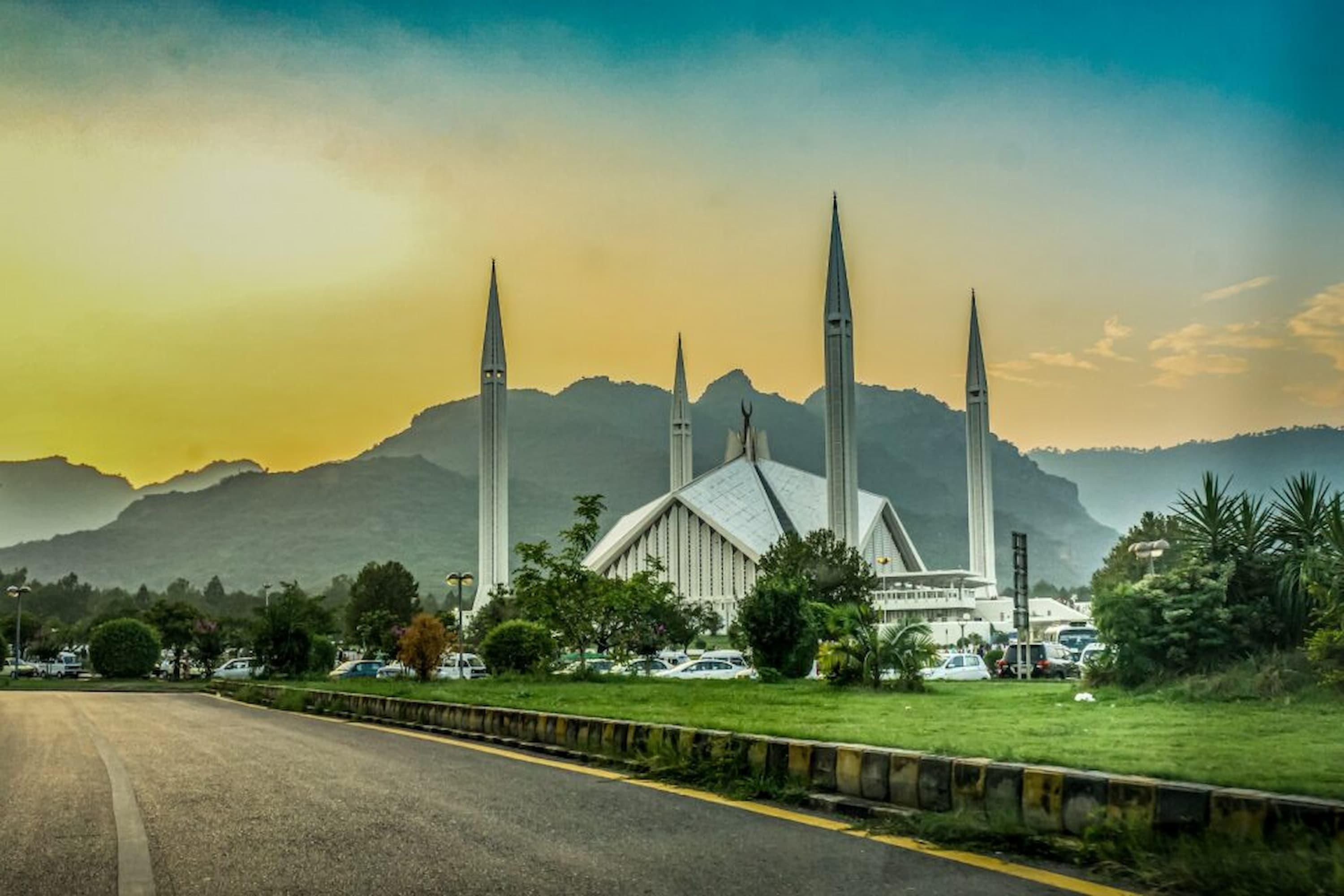 A mosque with tall minarets against a mountain backdrop at sunset, surrounded by greenery and parked cars.
