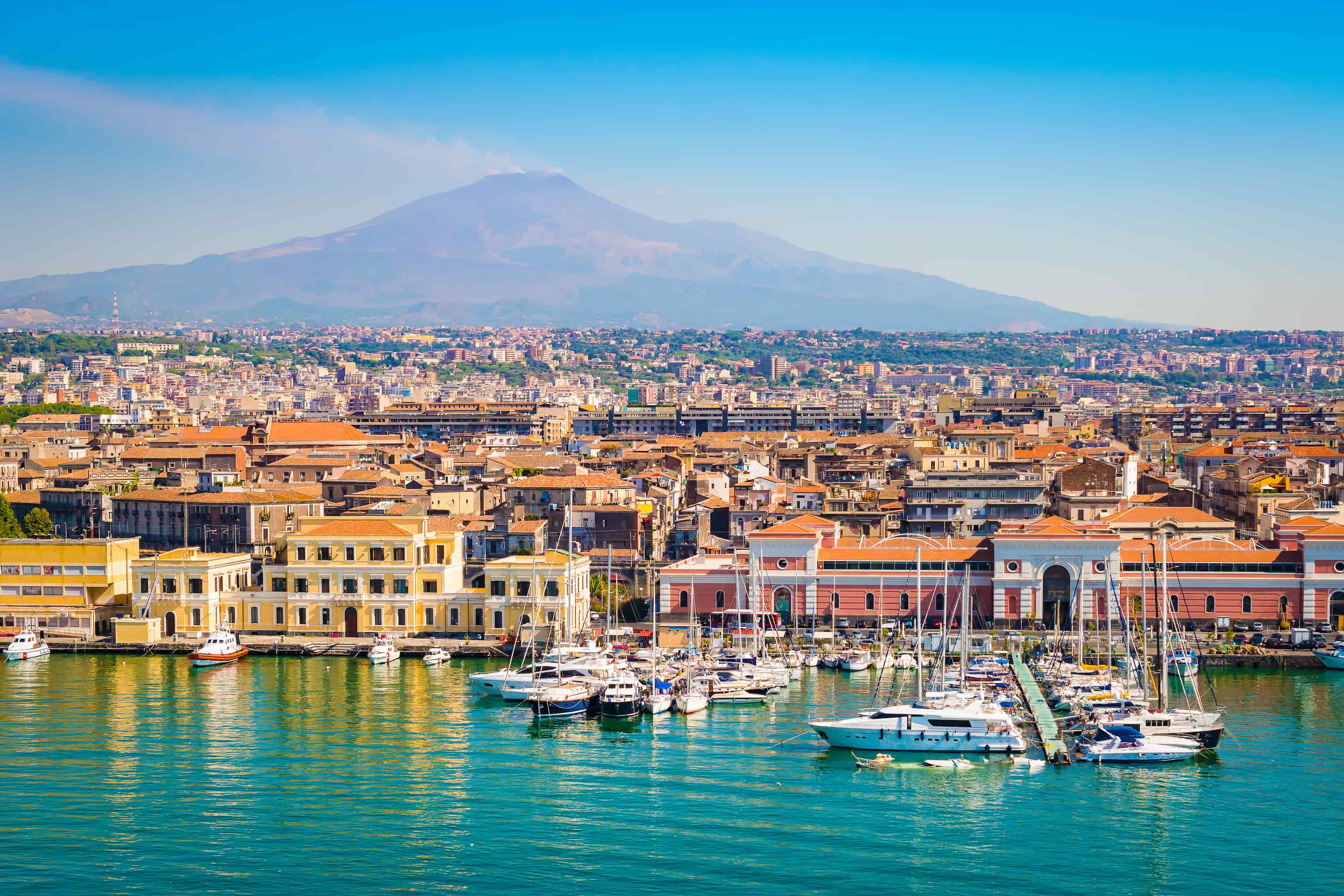 A seaside cityscape with a marina, colourful buildings, and a distant volcano under a clear blue sky.