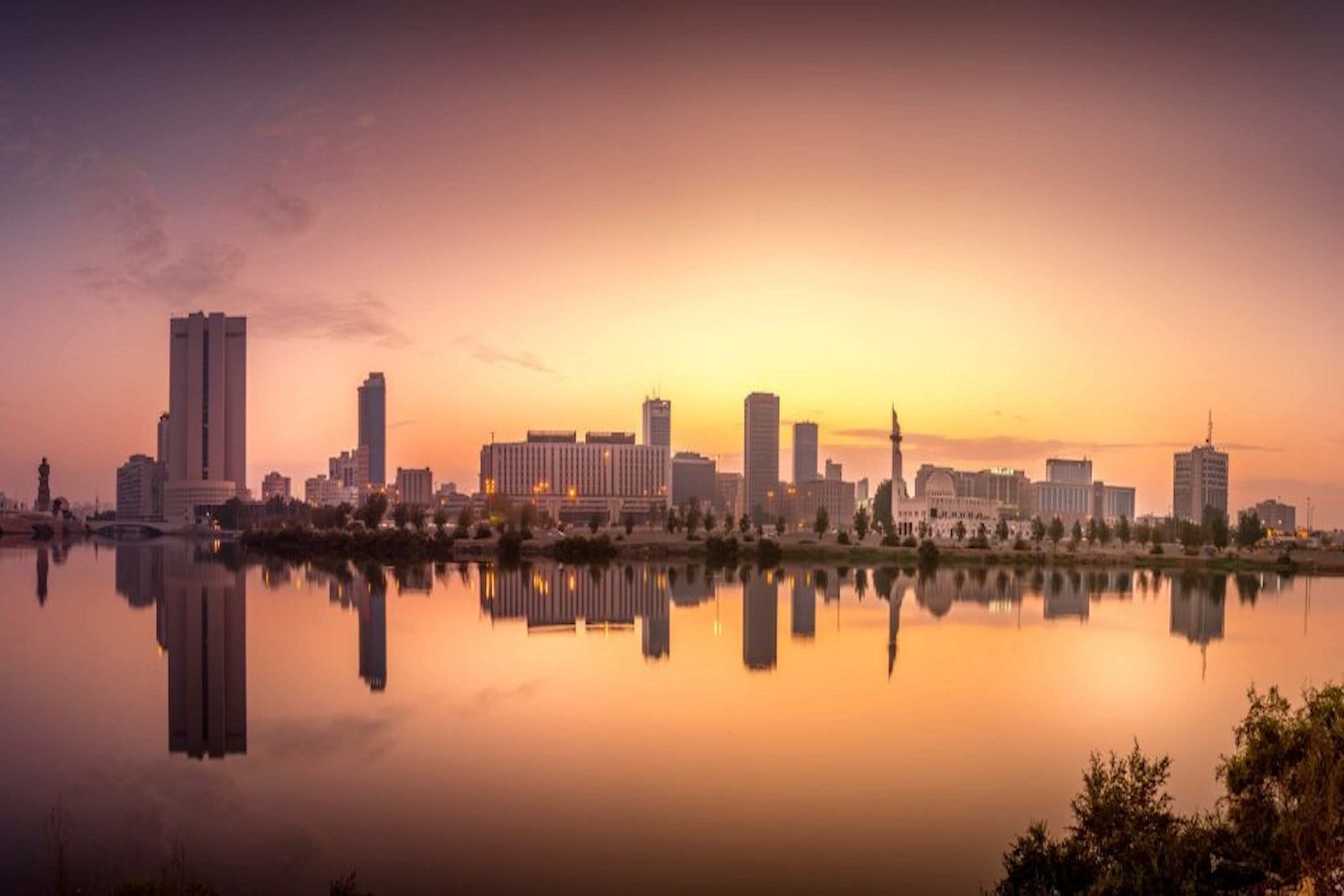 City skyline at sunset, reflected in a calm river, under a warm, glowing sky with silhouetted buildings and a prominent waterfront.
