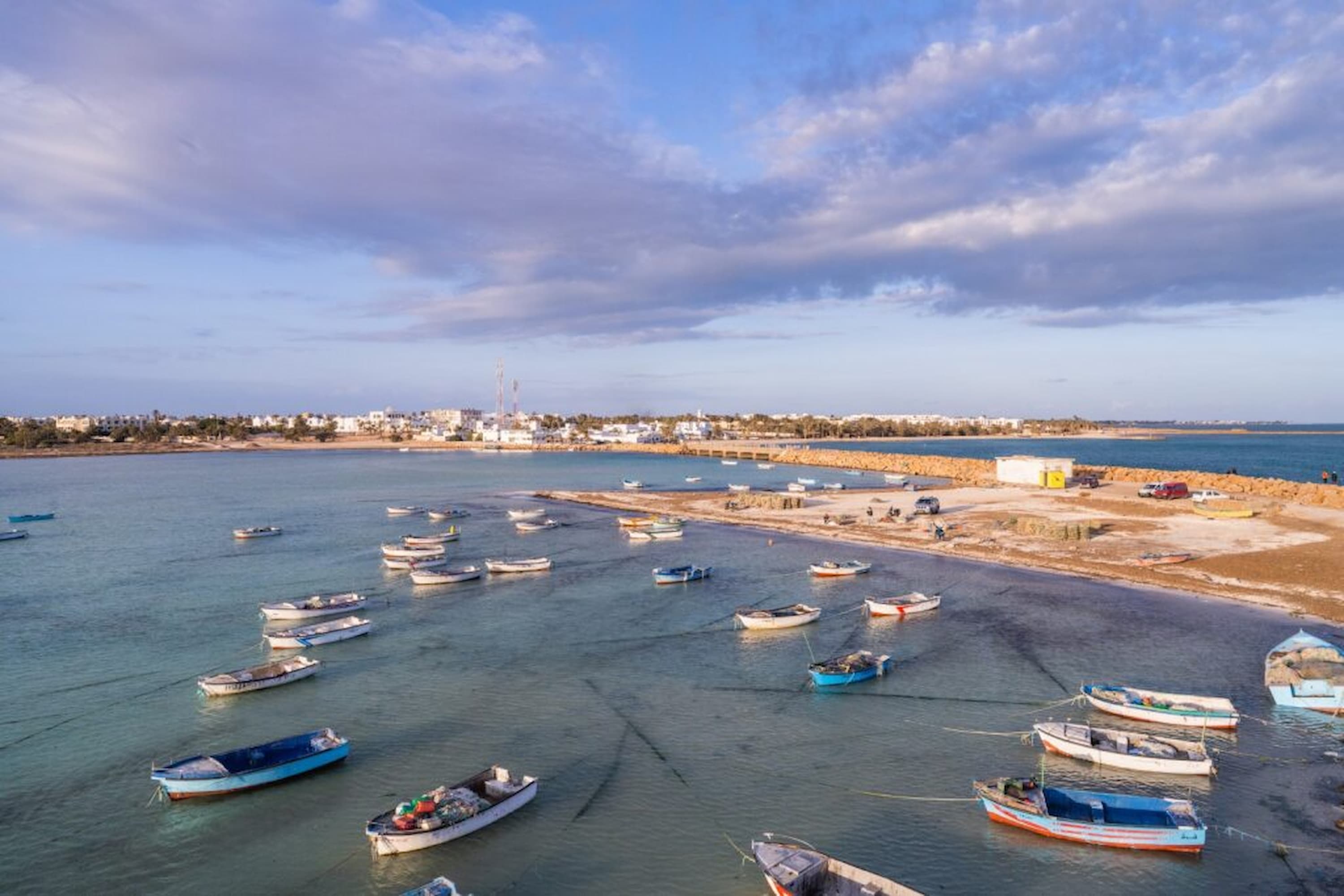 Aerial view of a calm coastal bay with numerous small boats, a sandy shore, and a distant town under a partly cloudy sky.