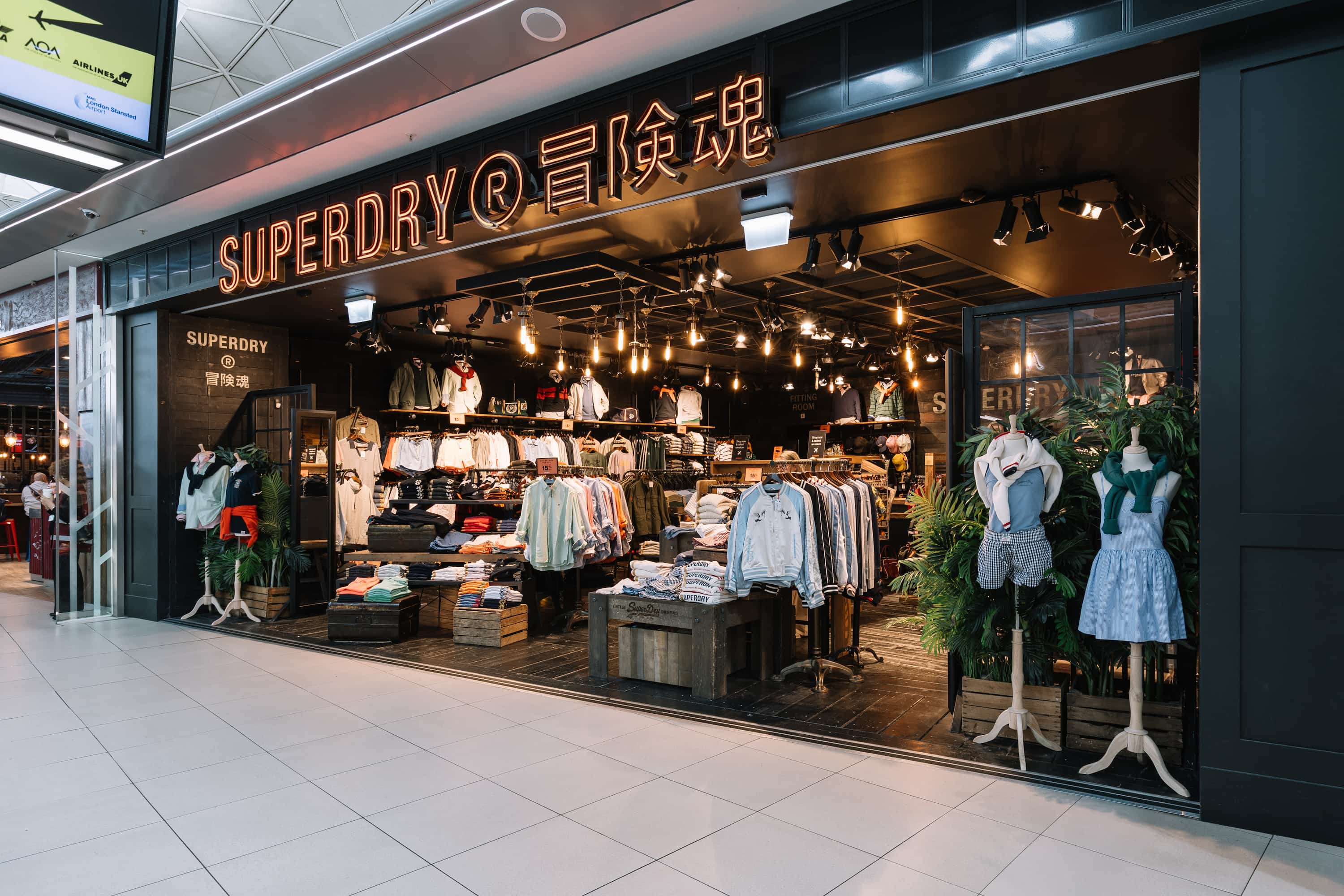 Front view of a Superdry store with clothing displays, racks, and mannequins, warmly lit in a shopping area.