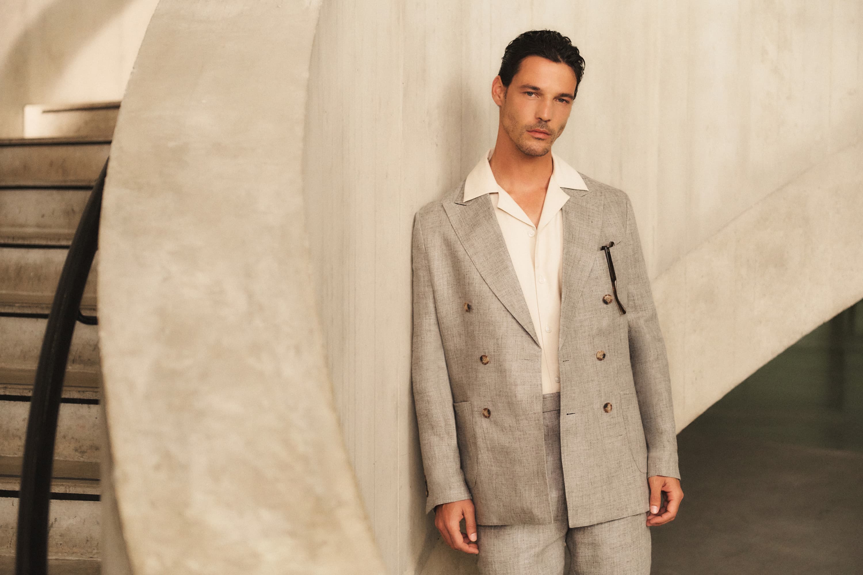Man in a light gray suit with a white shirt, leaning against a concrete wall near a staircase with a curved black railing.