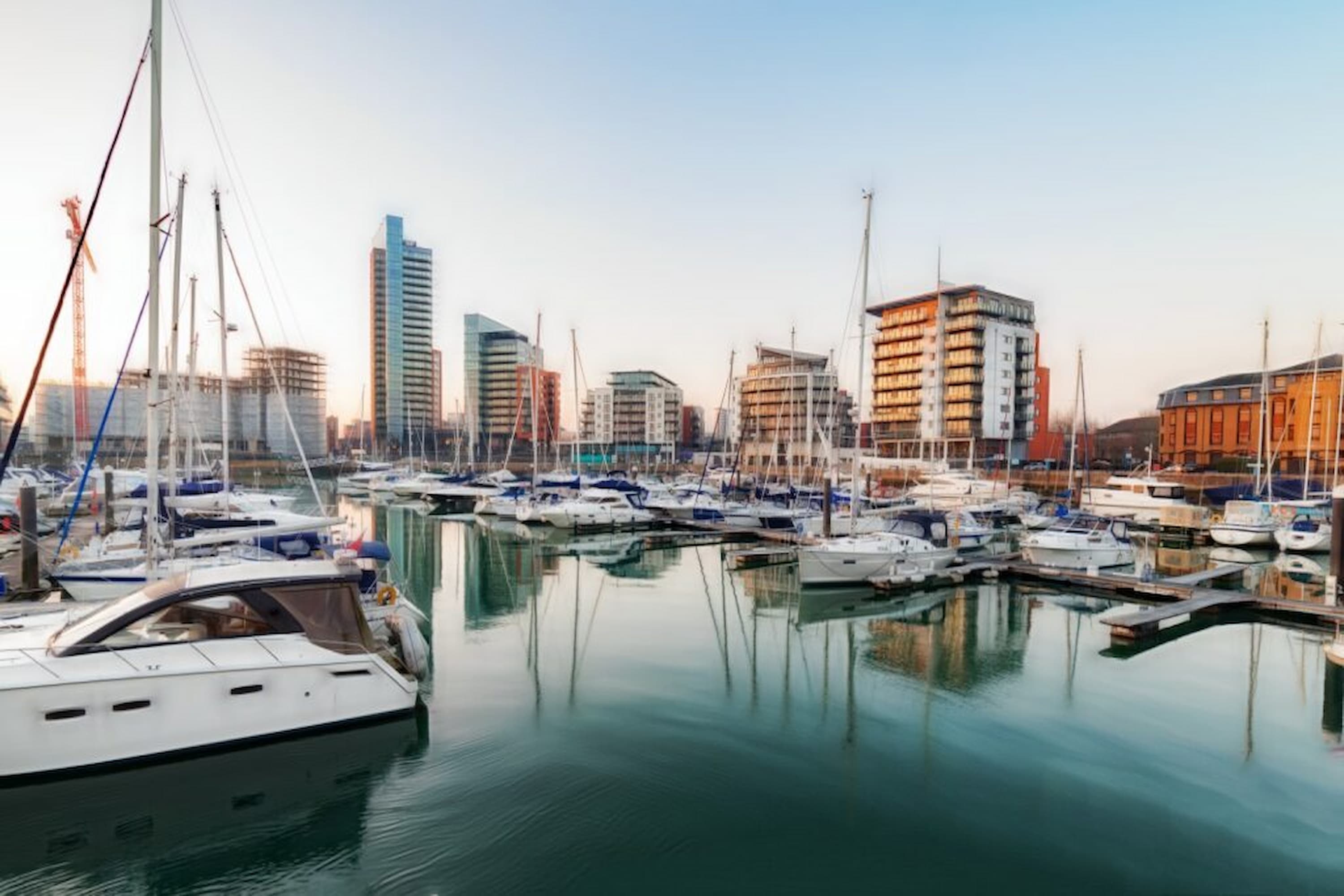 Marina with numerous sailboats and yachts docked by modern high-rise buildings under a clear sky at sunset.