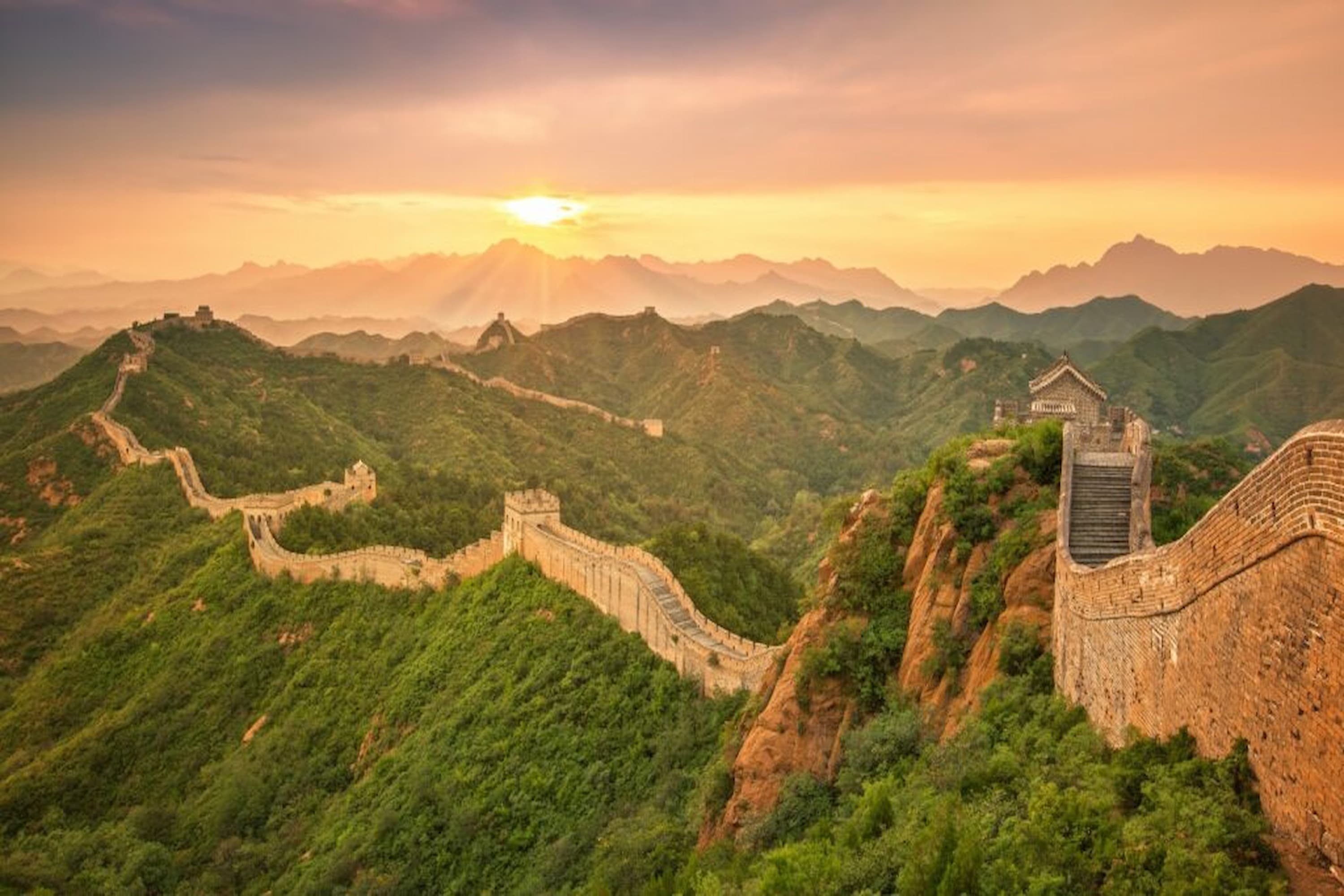 The Great Wall of China winding over lush green hills at sunset, with mountain silhouettes in the background.