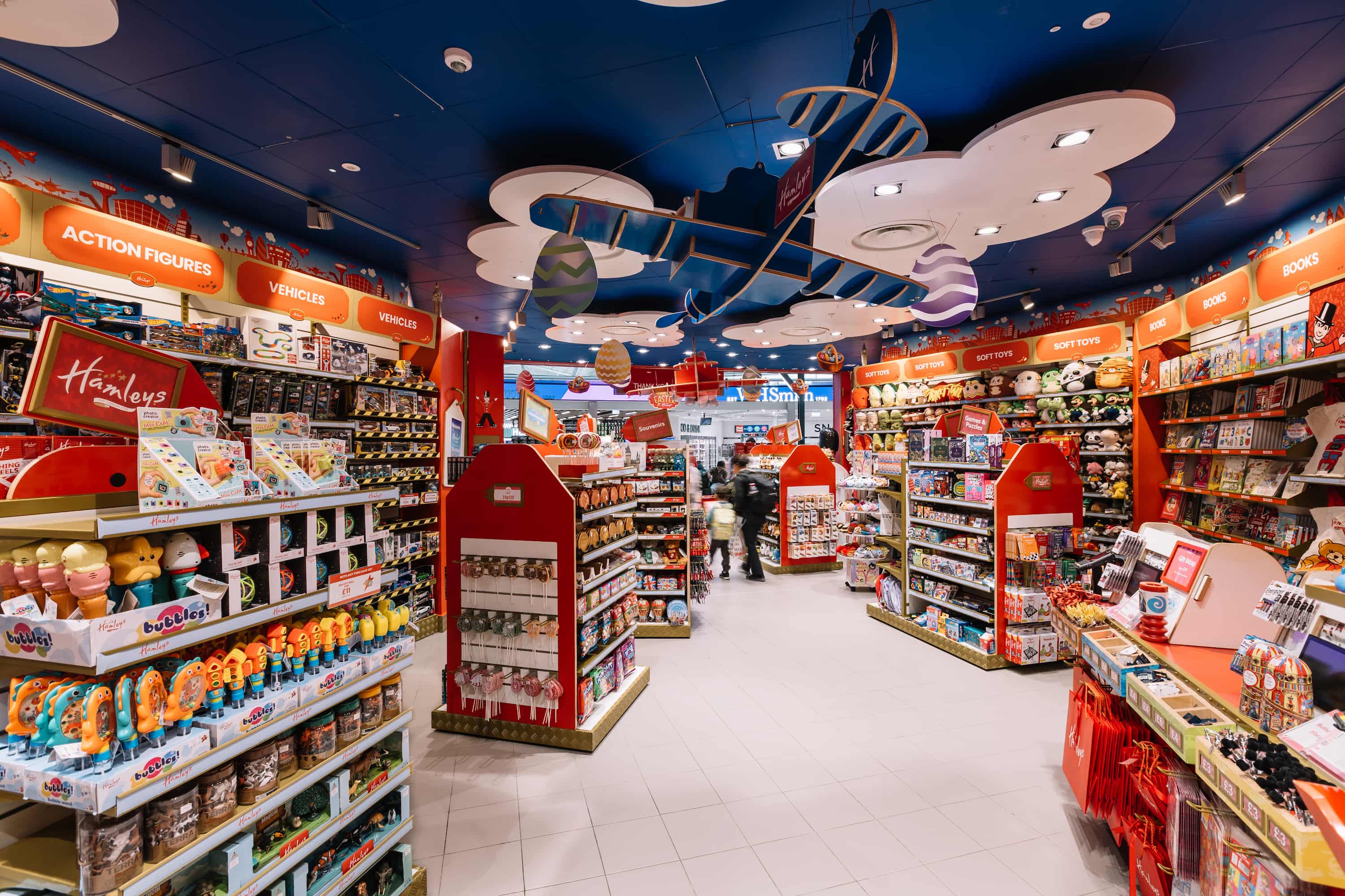 Colourful toy store interior with aisles of action figures, vehicles, and soft toys under a decorative ceiling featuring clouds and a plane.