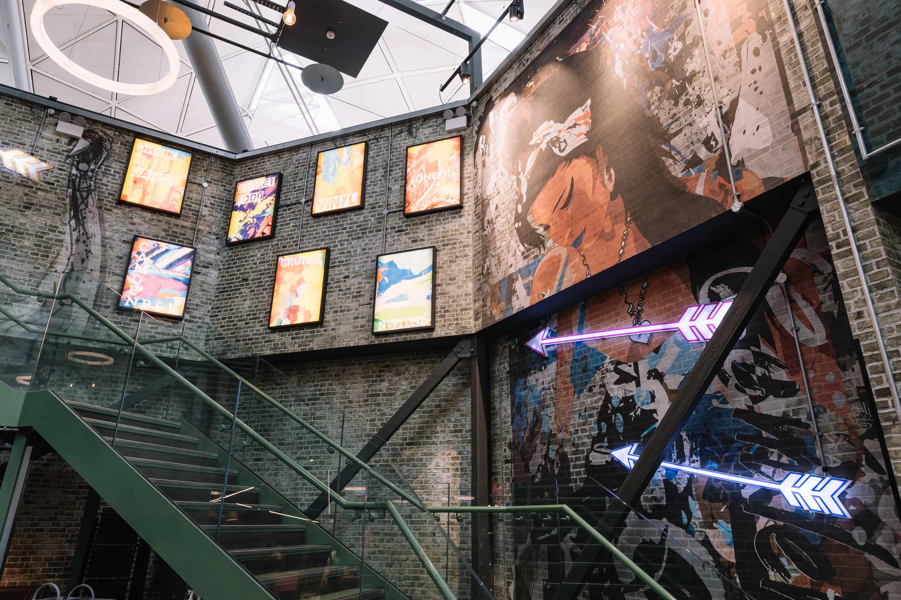 Modern interior with a brick wall featuring graffiti art, neon arrows, and vibrant framed posters. A glass staircase leads upwards.