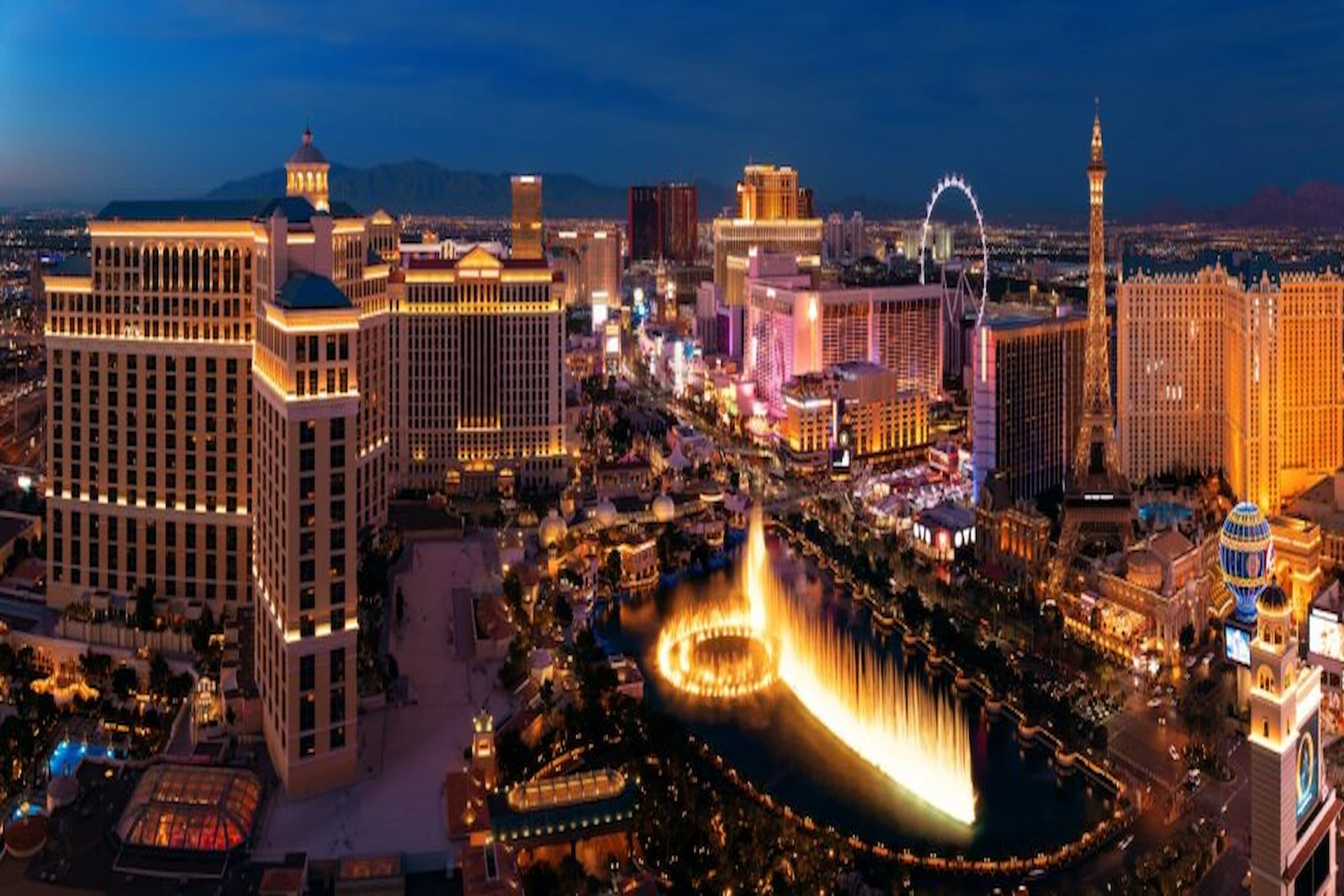 Panoramic view of the Las Vegas Strip at night, featuring illuminated hotels, casinos, fountains, and a large Ferris wheel against a dark sky.