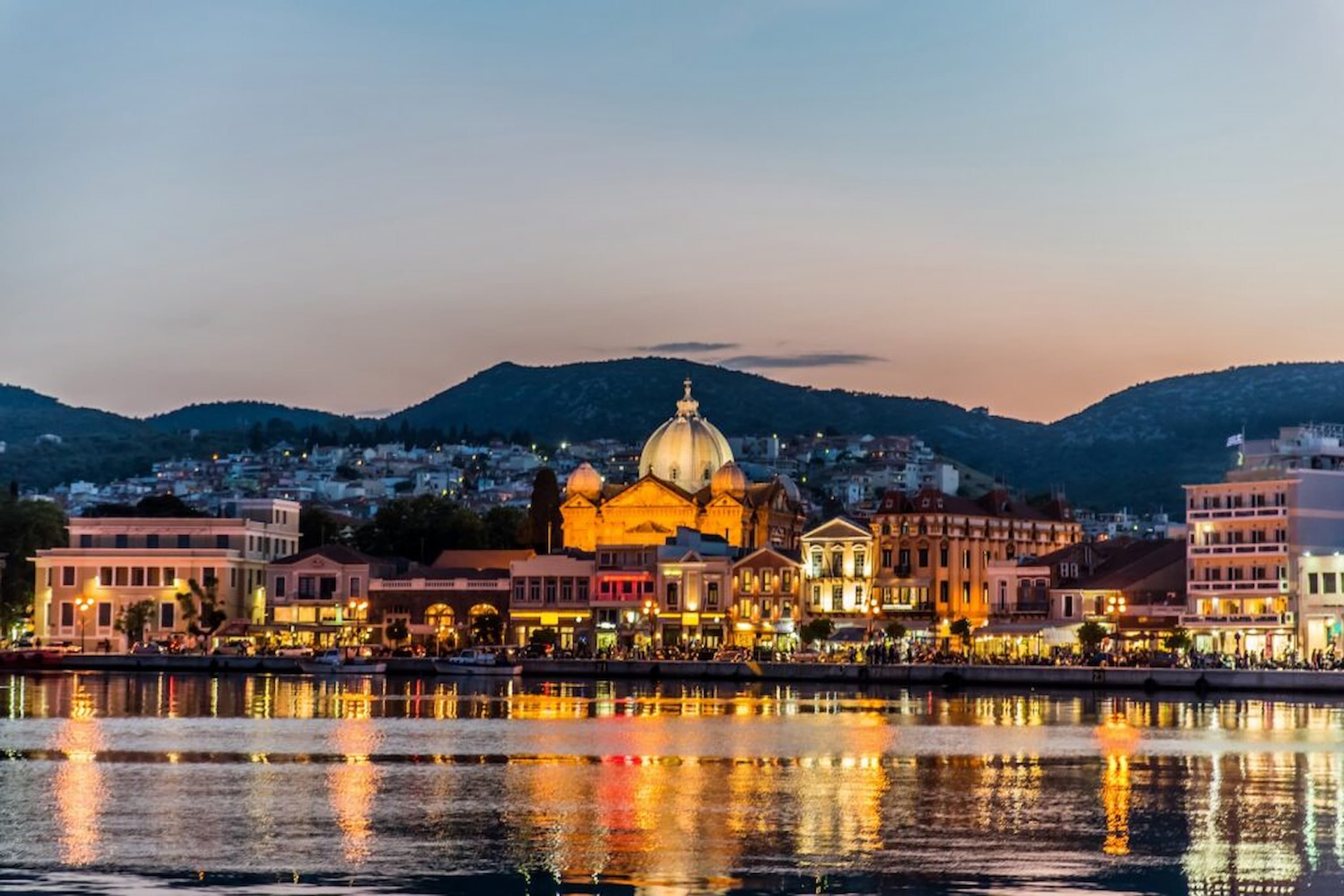 A scenic view of a coastal city at dusk, featuring illuminated buildings and a prominent domed structure reflected in the water.