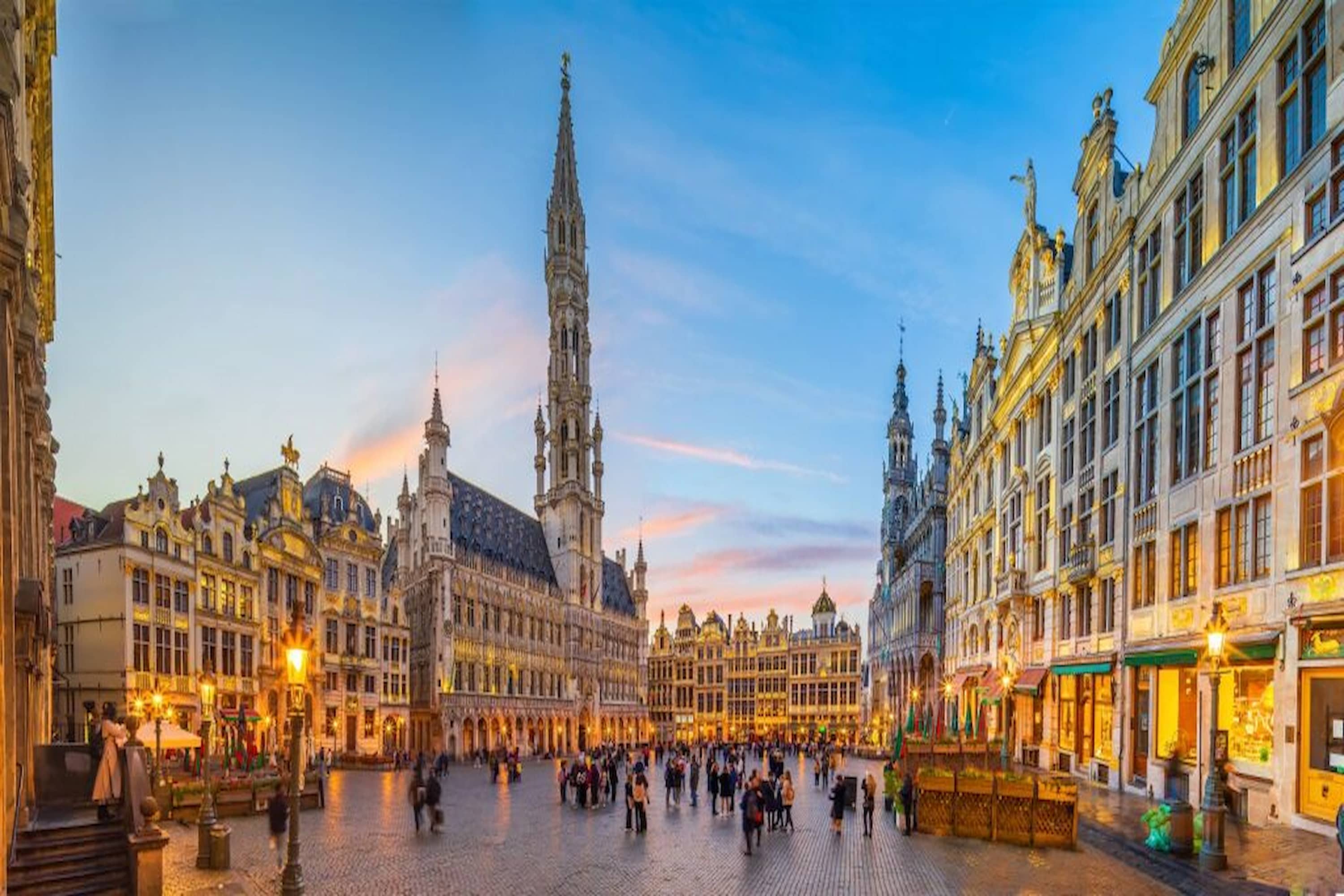People gather in Brussels' Grand Place, surrounded by ornate, historic buildings under a twilight sky.