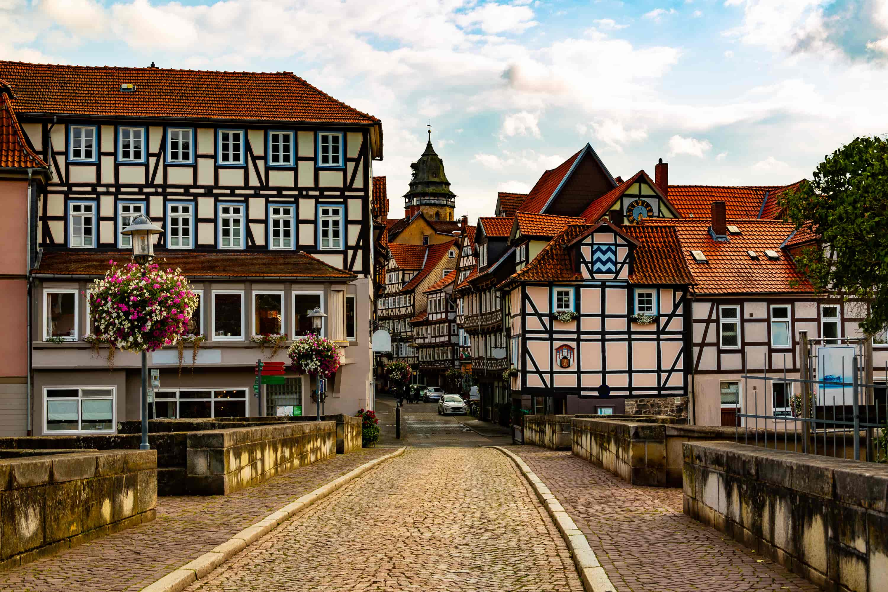 Cobblestone street leading to traditional half-timbered houses with red roofs, adorned with hanging flowers and a tower in the background.