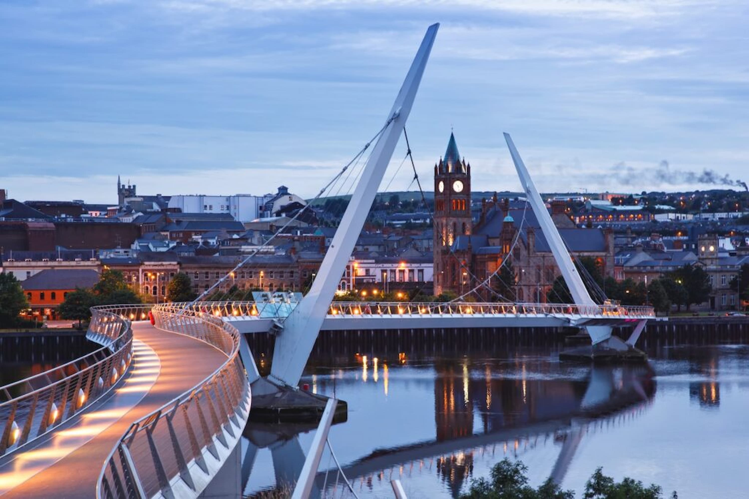Peace Bridge in Derry, Northern Ireland, illuminated at dusk with cityscape and river reflections, featuring distant historical architecture.