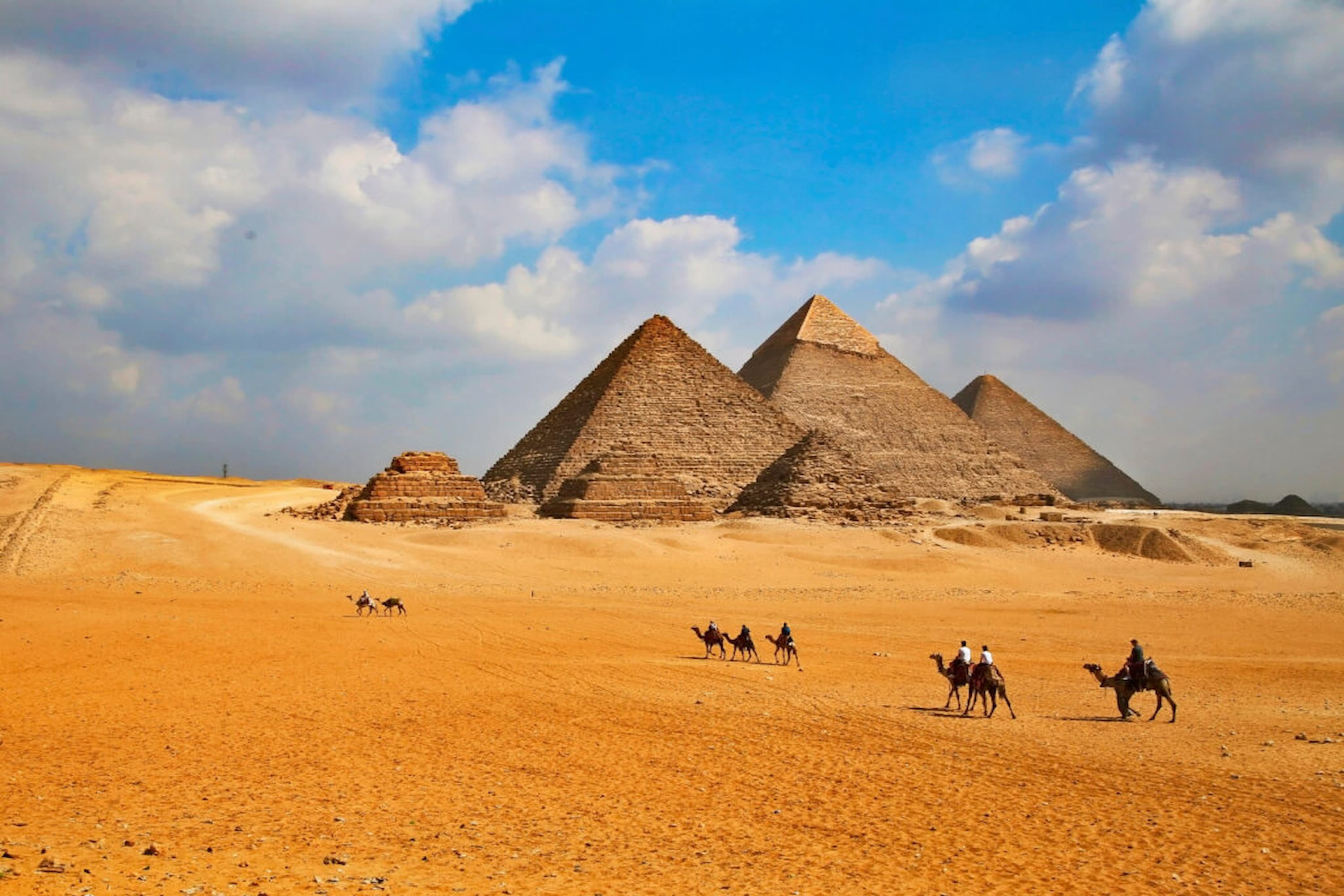 Camel riders approaching the Pyramids of Giza under a partly cloudy sky, with desert sand in the foreground.