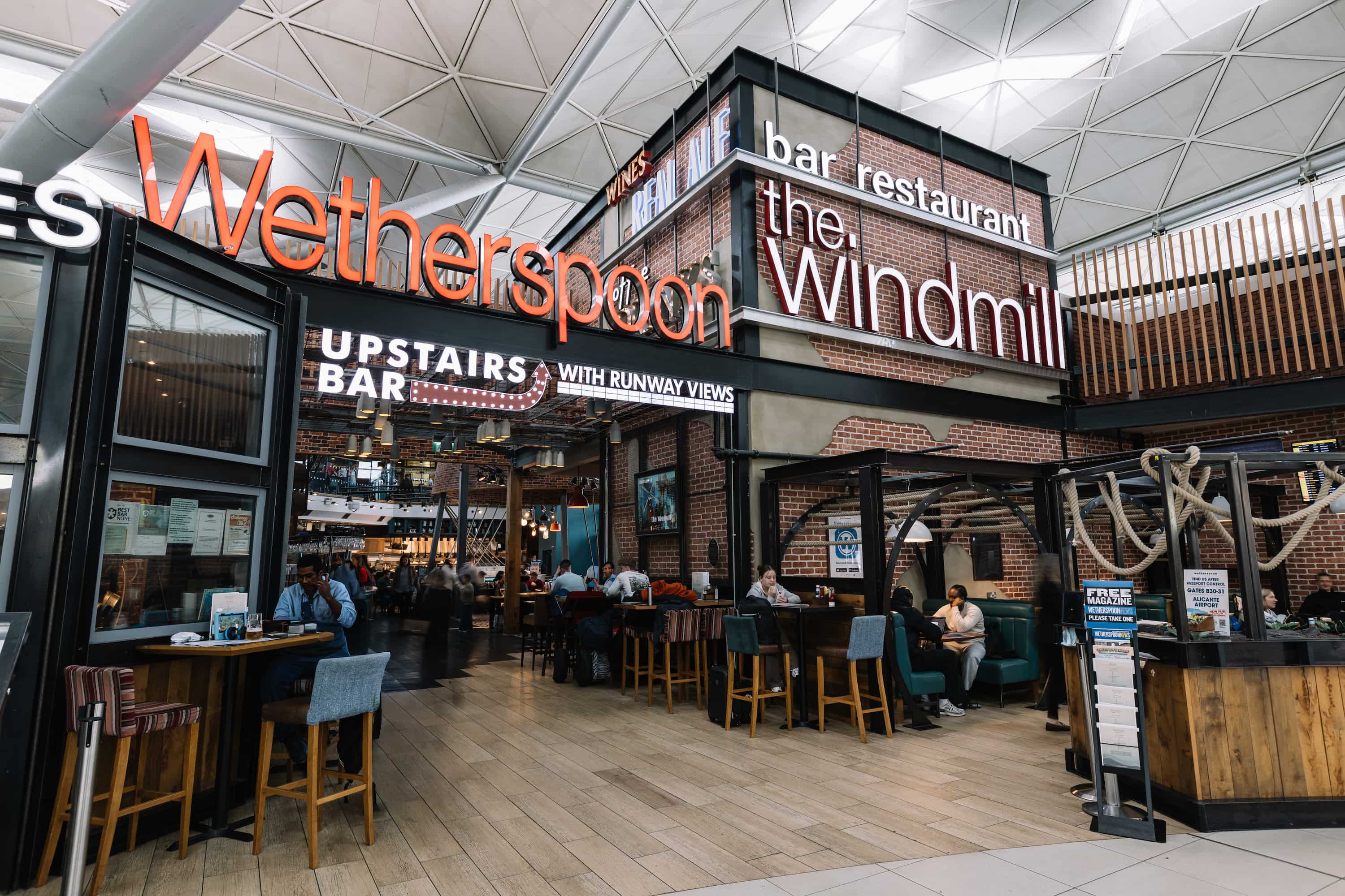 Wetherspoon's The Windmill bar and restaurant in an airport, with people dining and signs highlighting an upstairs bar with runway views.