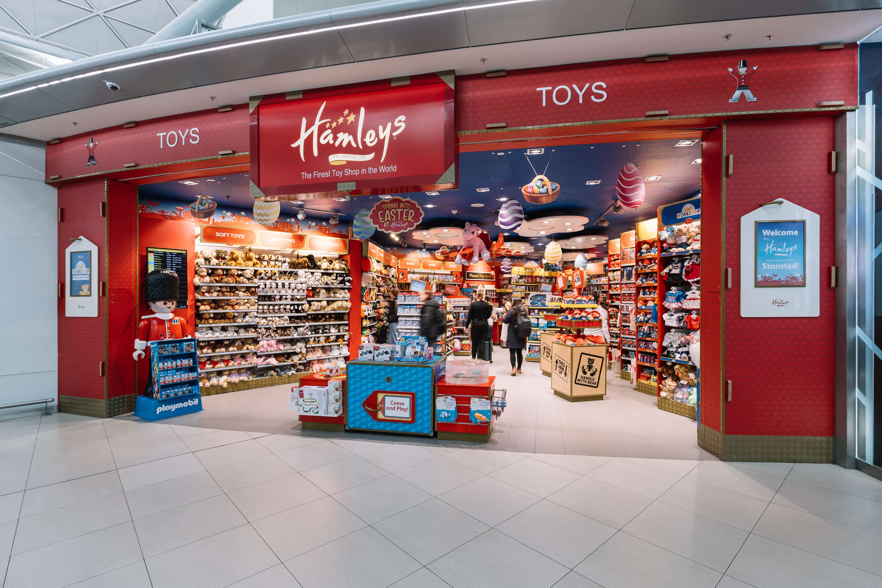 Entrance to a brightly lit toy store with colourful displays, stuffed animals, and an eye-catching sign overhead. Shoppers inside browsing.