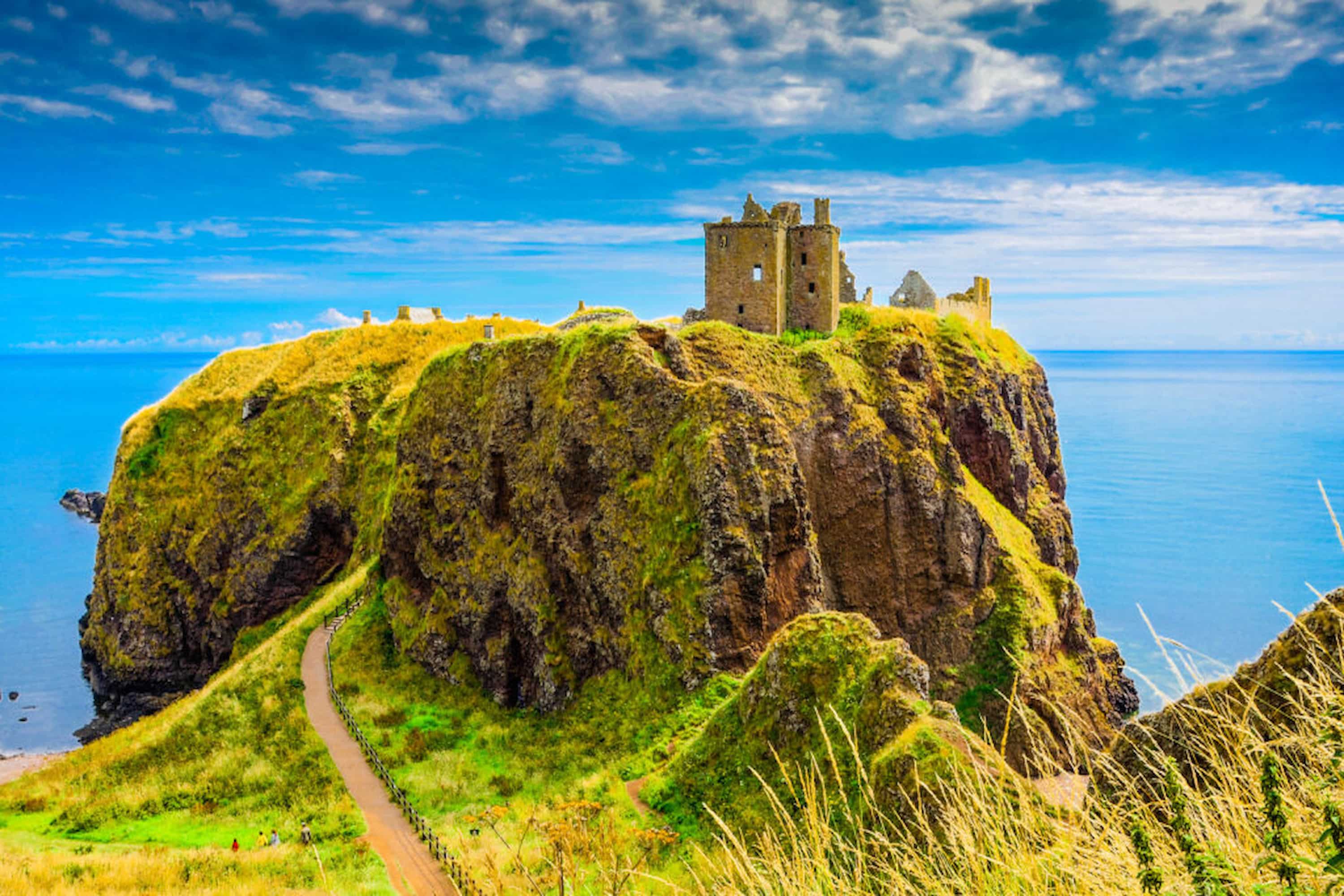 Ancient stone castle ruins atop a steep grassy cliff overlooking a calm blue sea under a partly cloudy sky