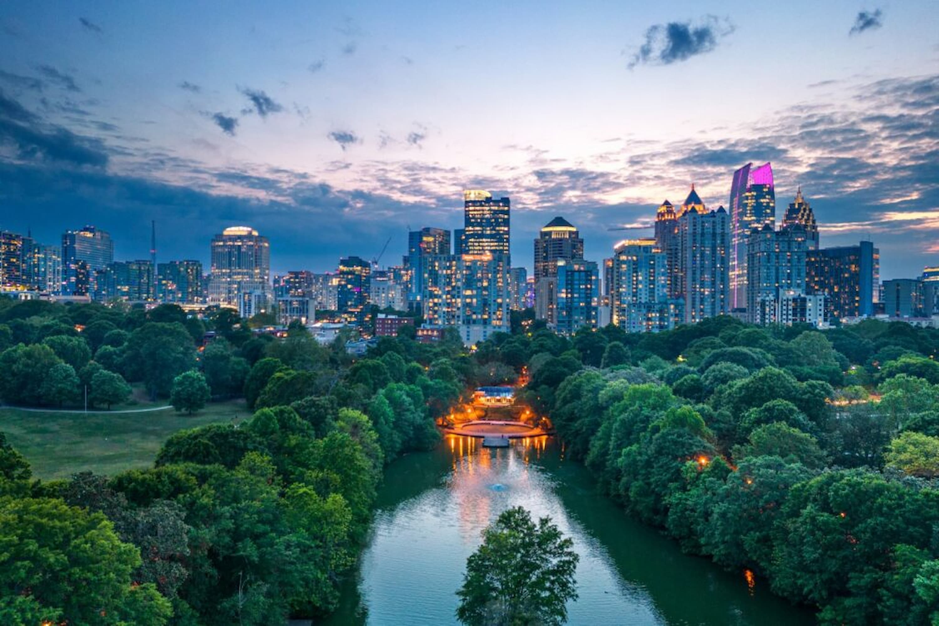 A serene cityscape at dusk with a river and lush trees in the foreground