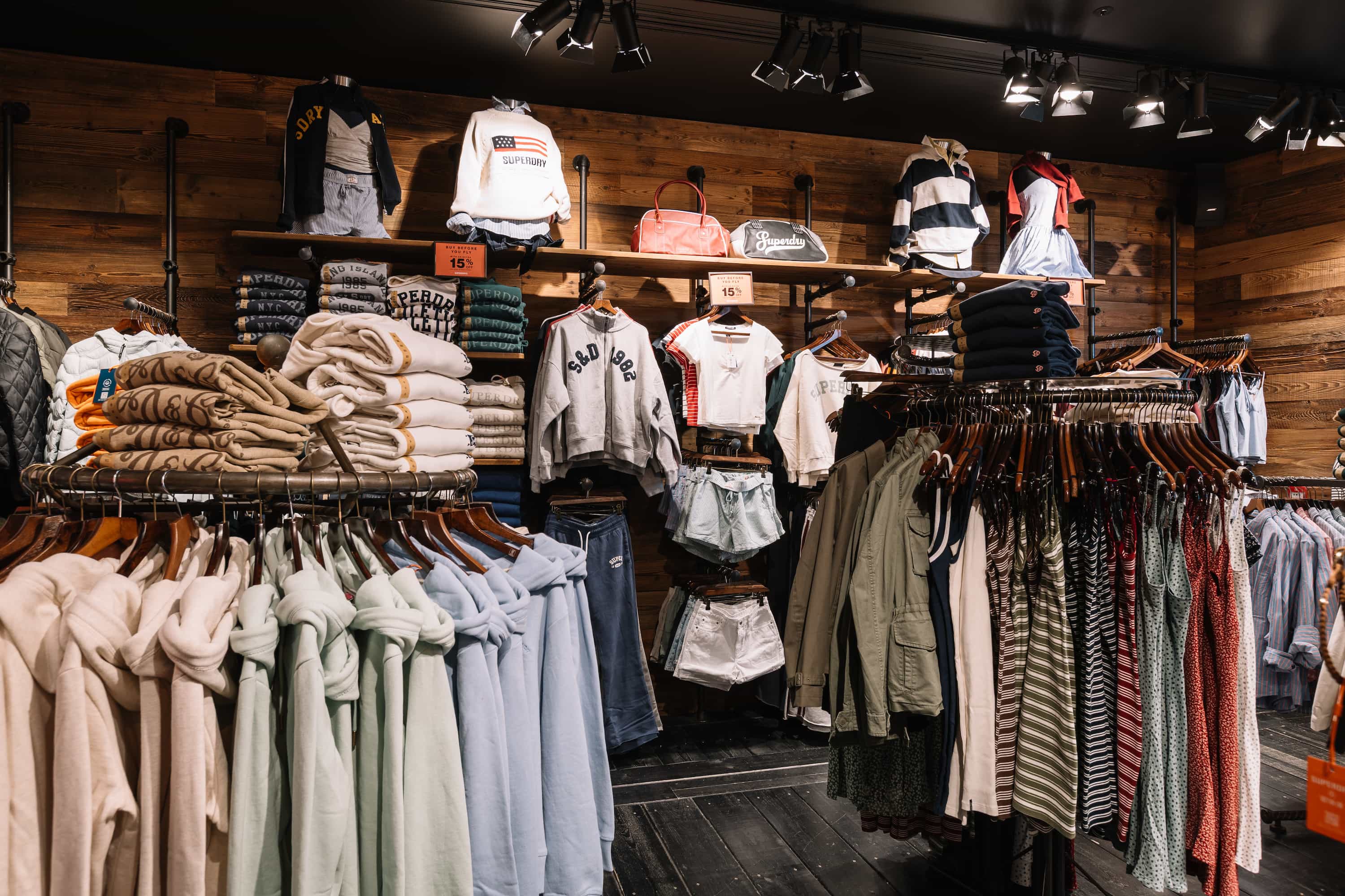 Clothing store interior with hoodies, sweaters, shorts, and jackets displayed on racks against a wooden wall, under bright lighting.