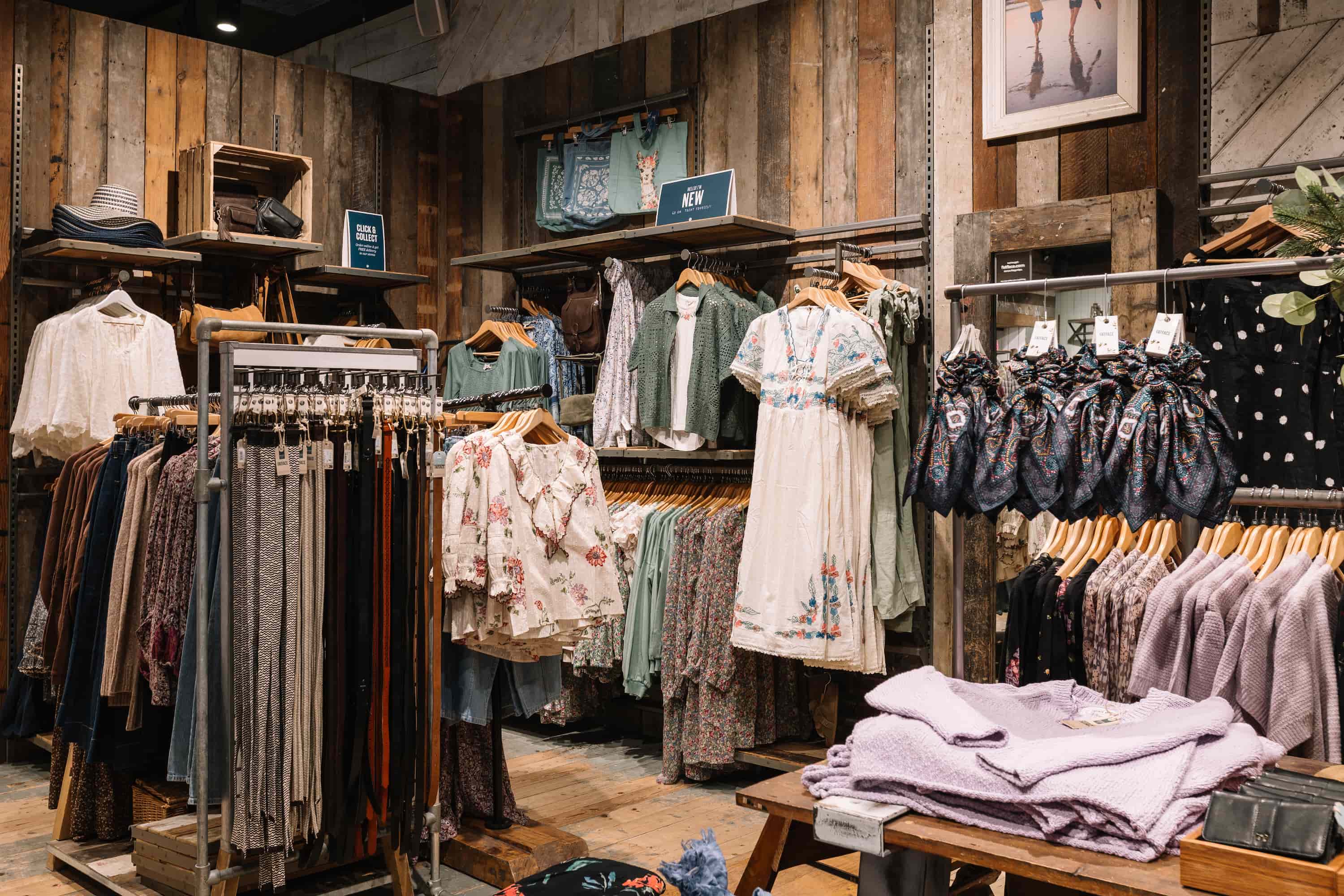 Clothing store interior with floral and patterned dresses, scarves, and display of pastel sweaters on rustic wooden shelves.