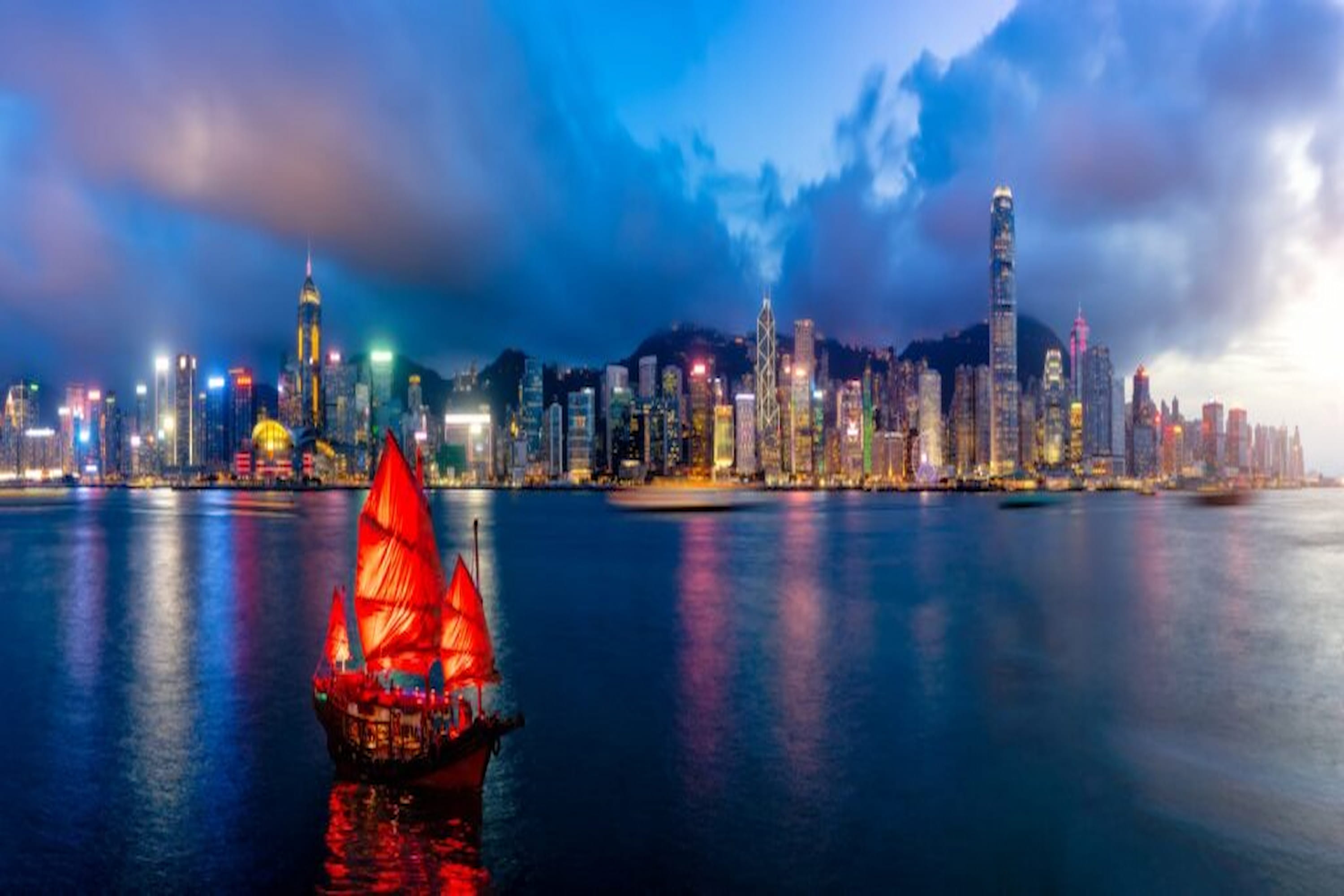 A traditional red-sailed junk boat on calm water with a vibrant city skyline illuminated at dusk in the background.