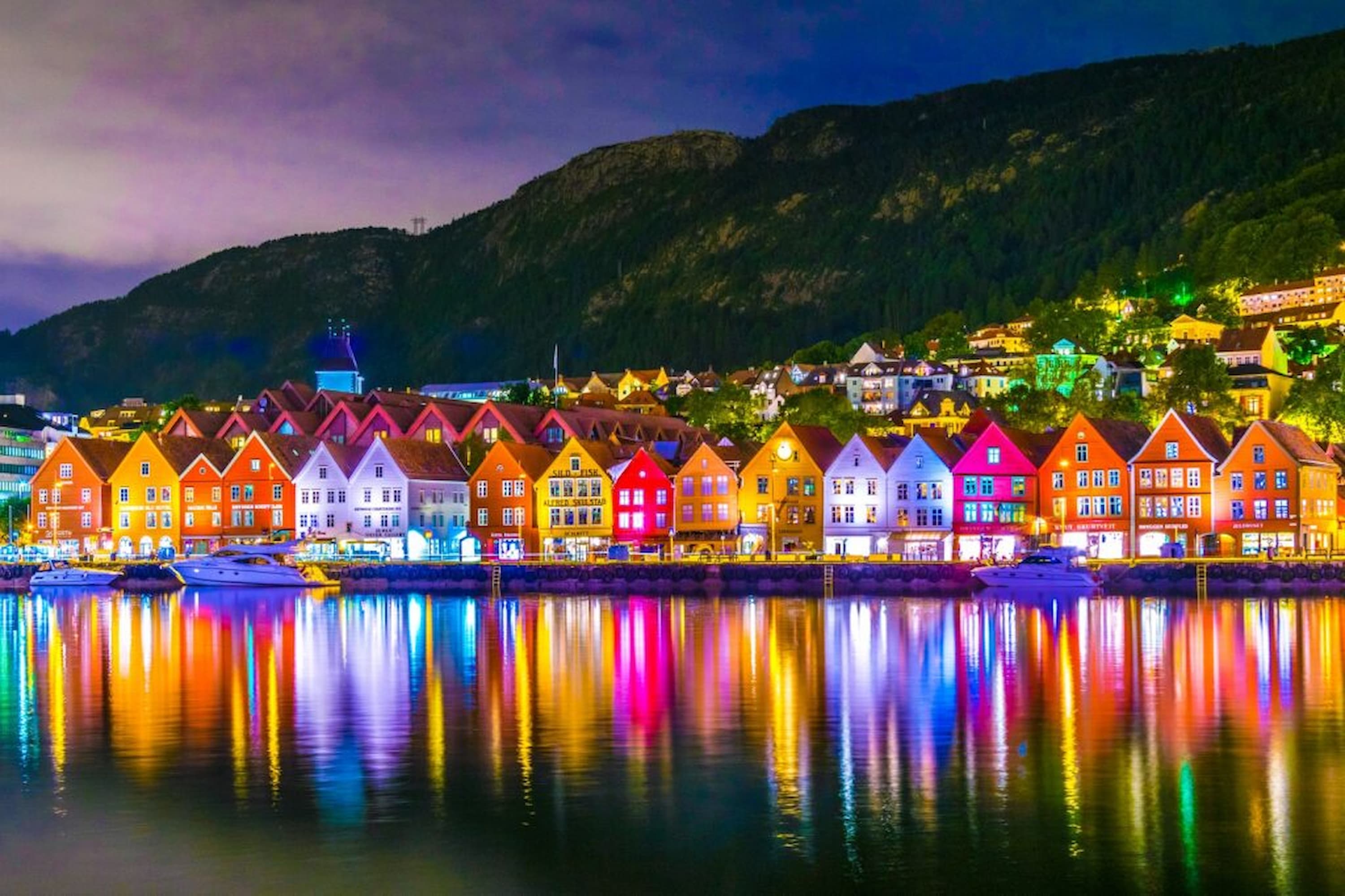 Colourful wooden buildings along a waterfront at night, reflecting vibrant lights on the water, with a forested hillside in the background.
