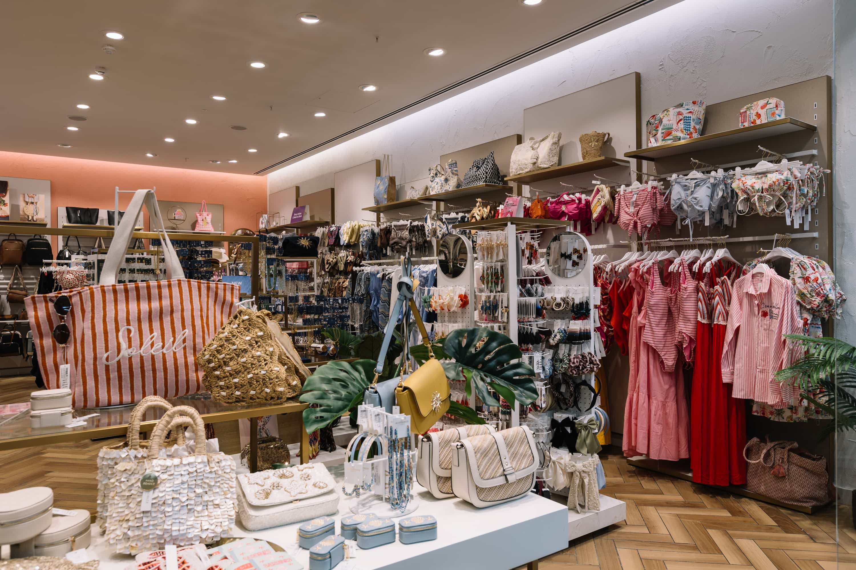 Clothing store interior featuring racks of colourful bags, dresses, jewellery, and accessories on display, with bright lighting and wooden flooring.