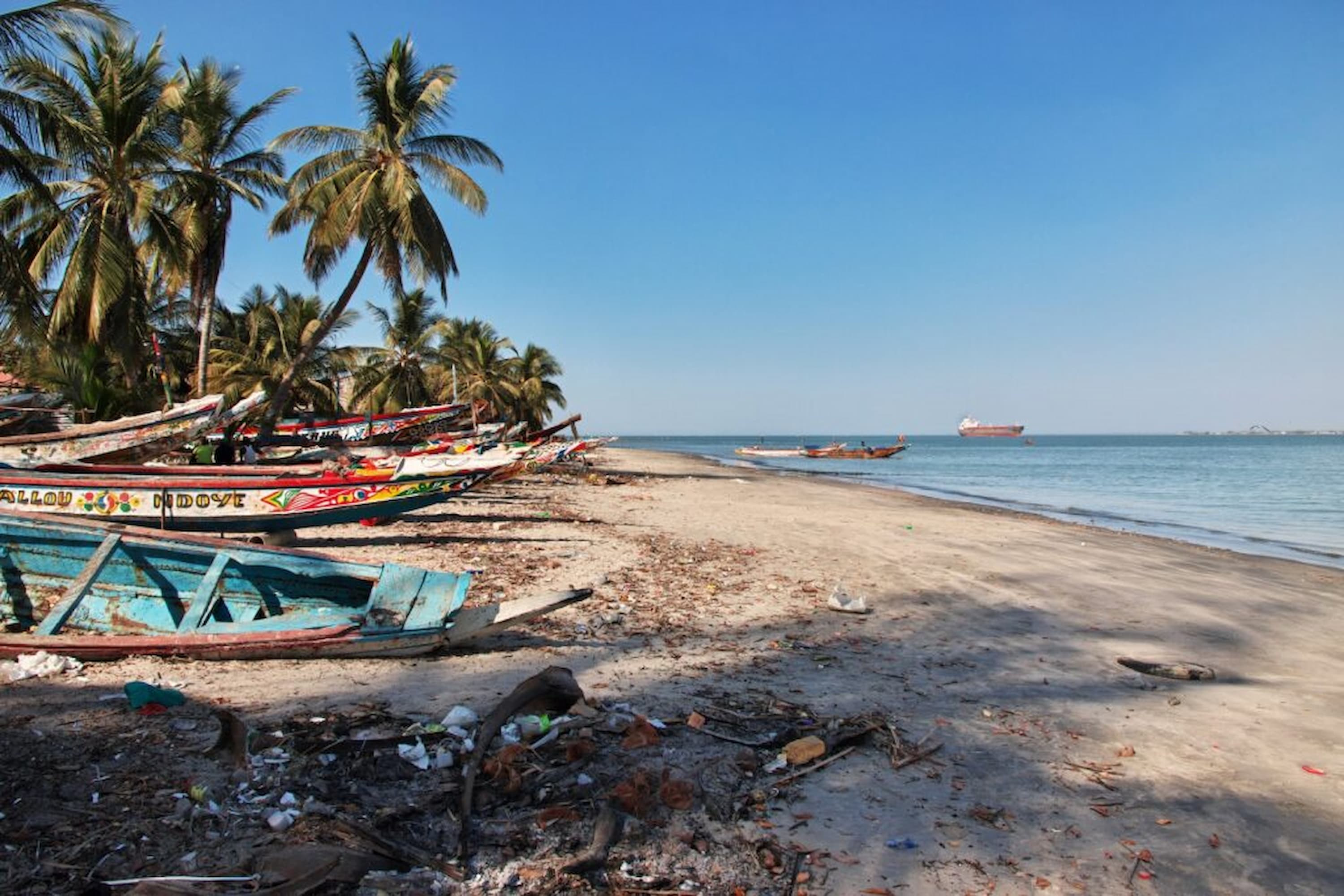 Colourful fishing boats on a sandy beach with palm trees, debris, and a ship in the distance under a clear blue sky.