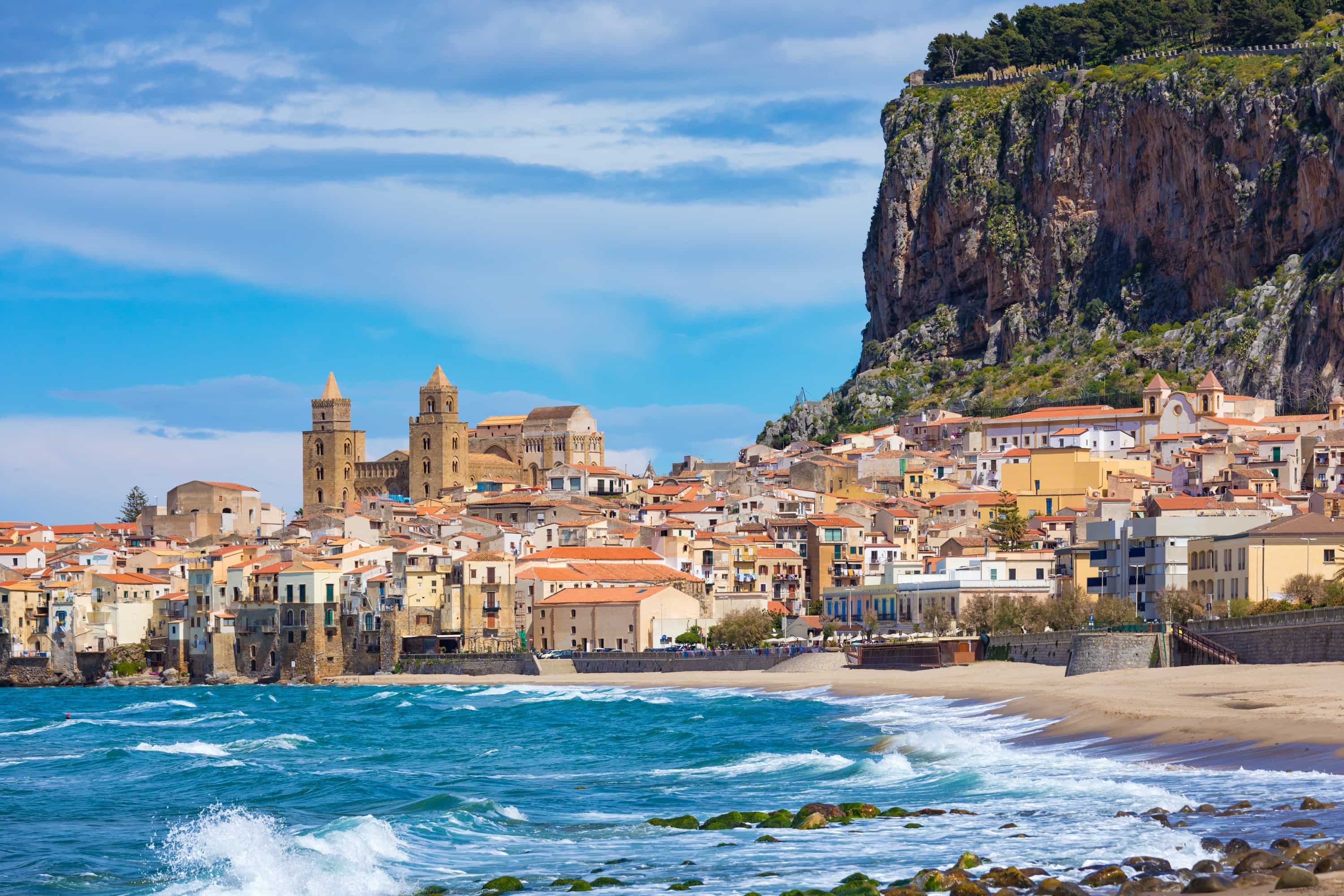 A picturesque coastal town with historic buildings and a cathedral, set against a rocky cliff and a clear blue sky, with waves crashing on the shore.