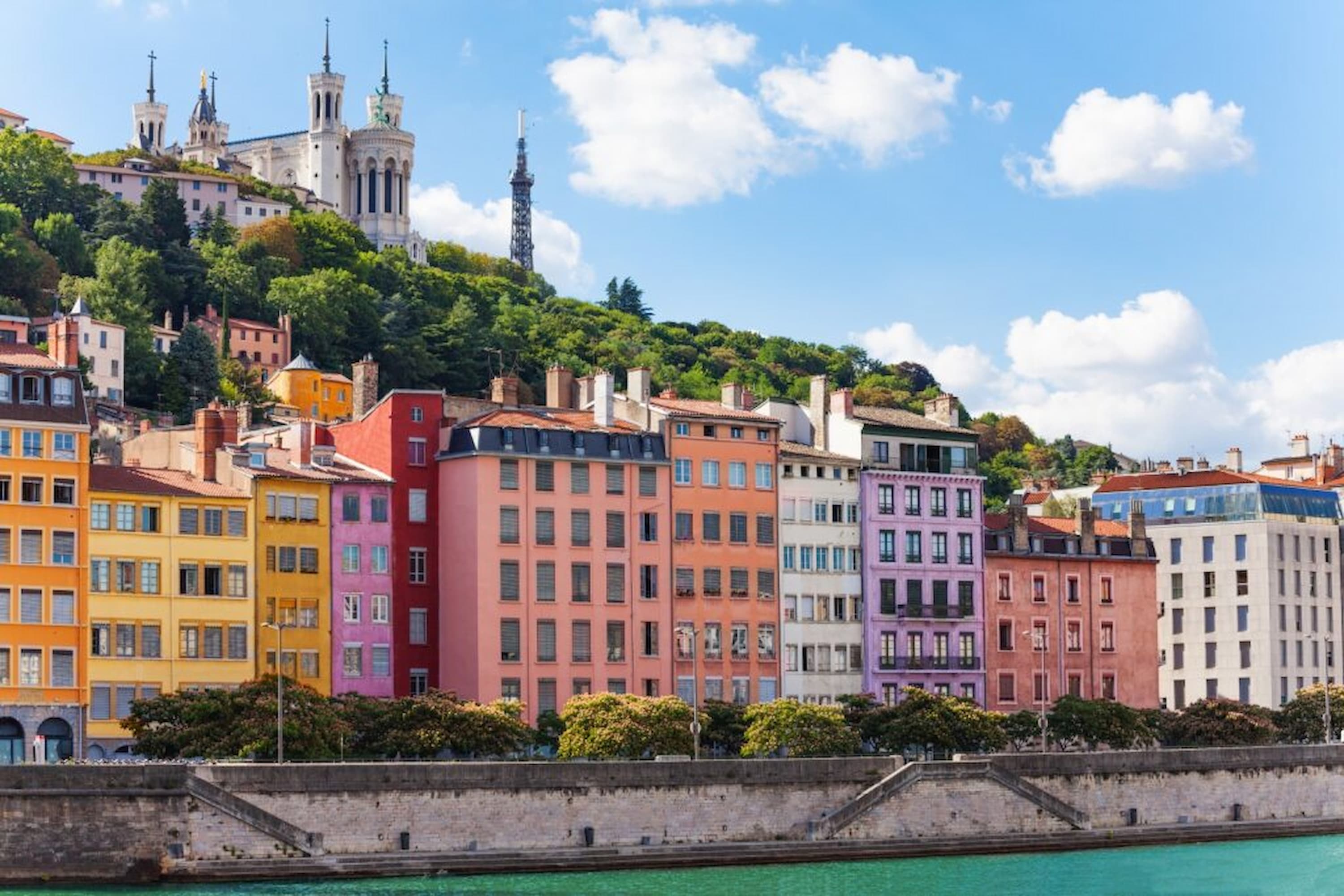 Colourful buildings line a riverbank in Lyon, France, with the Basilica of Notre-Dame de Fourvière on a hill under a blue sky with clouds.