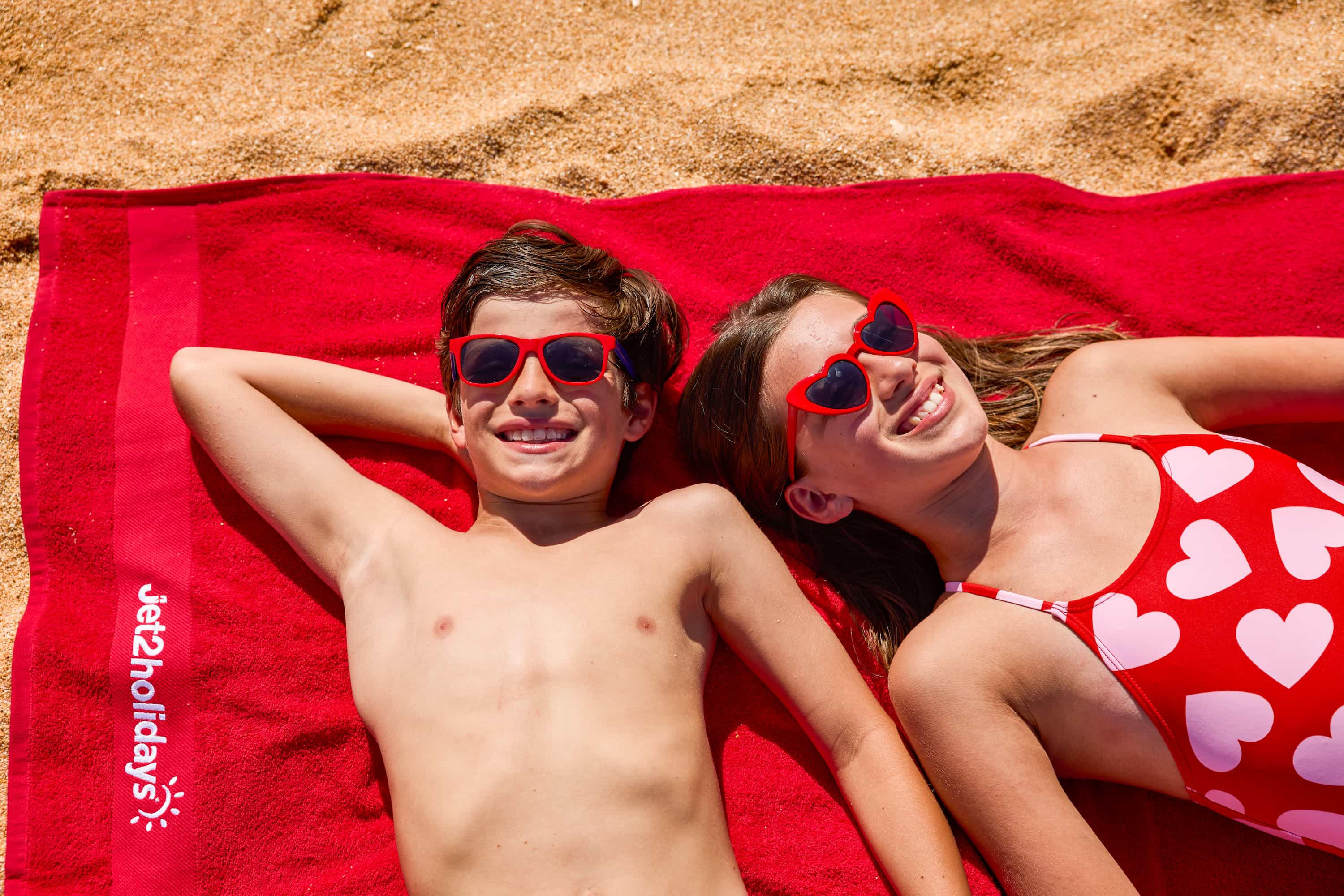 Two kids wearing red sunglasses sunbathe on a red beach towel with heart designs, smiling and enjoying the sunny day.