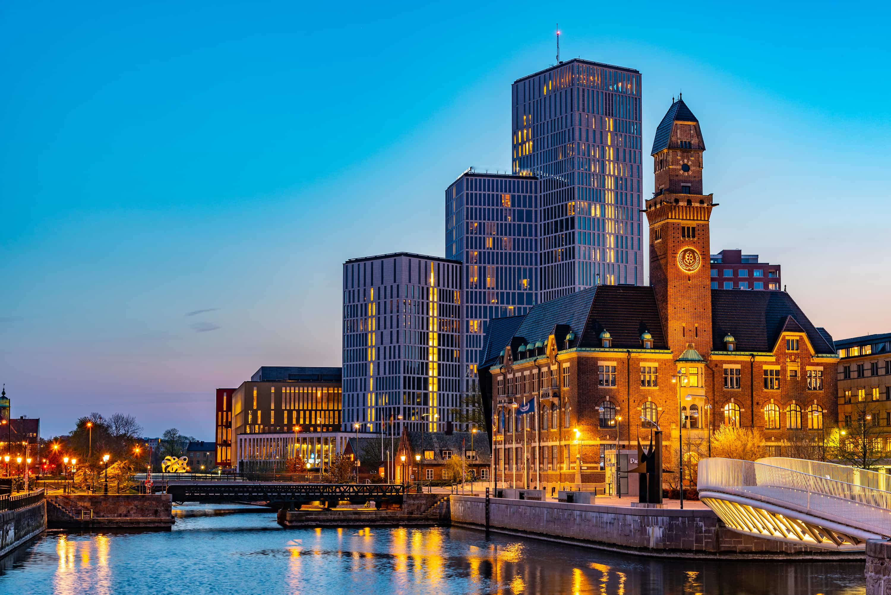 Cityscape at dusk with illuminated modern skyscrapers and historic buildings reflected in a canal.