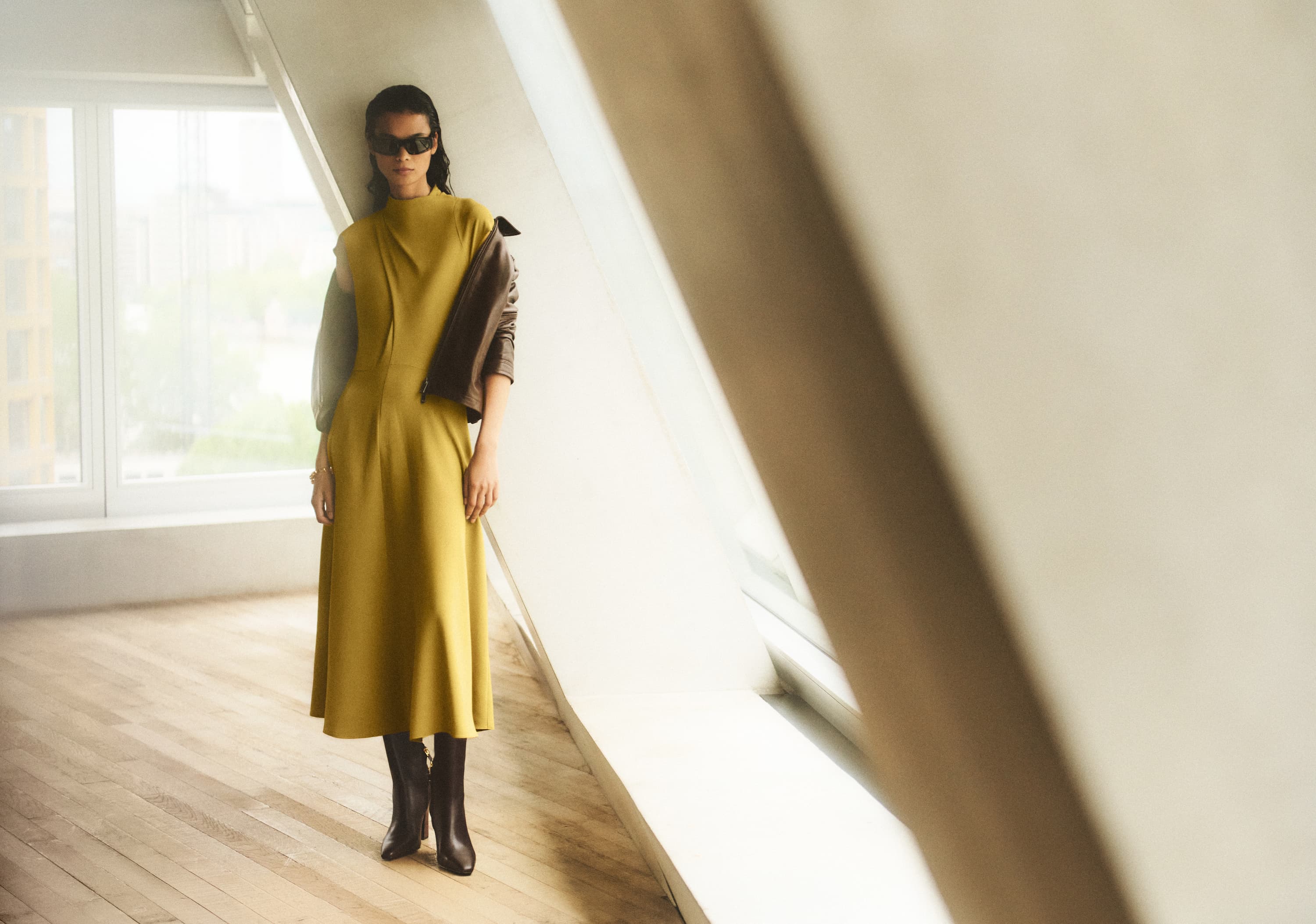 Person in a long mustard dress and sunglasses, wearing boots, stands in a sunlit room with angled windows and wooden floors.