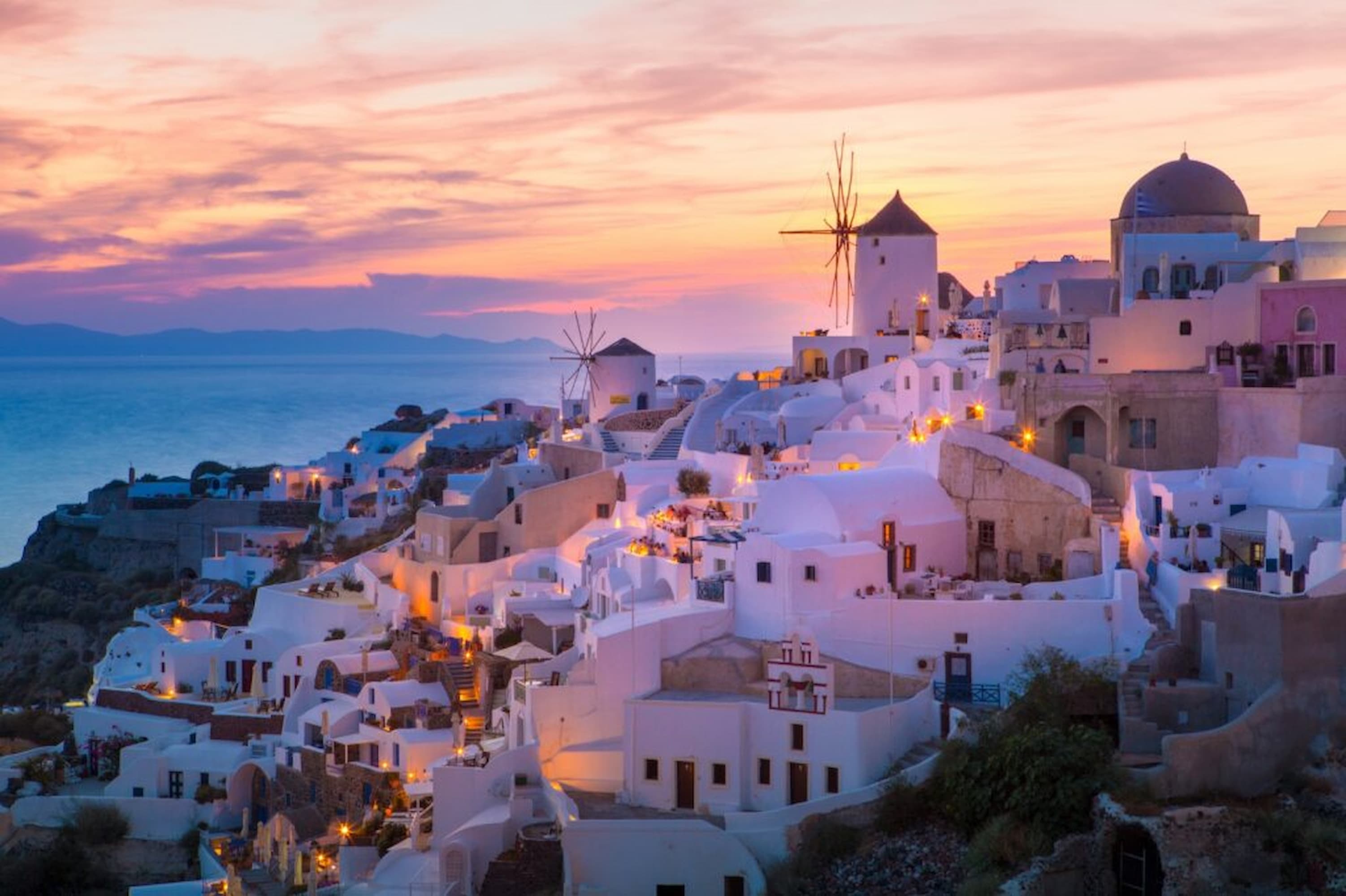 Sunset view of Santorini, Greece, with white buildings, windmills, and the sea in the background, all bathed in warm evening light.