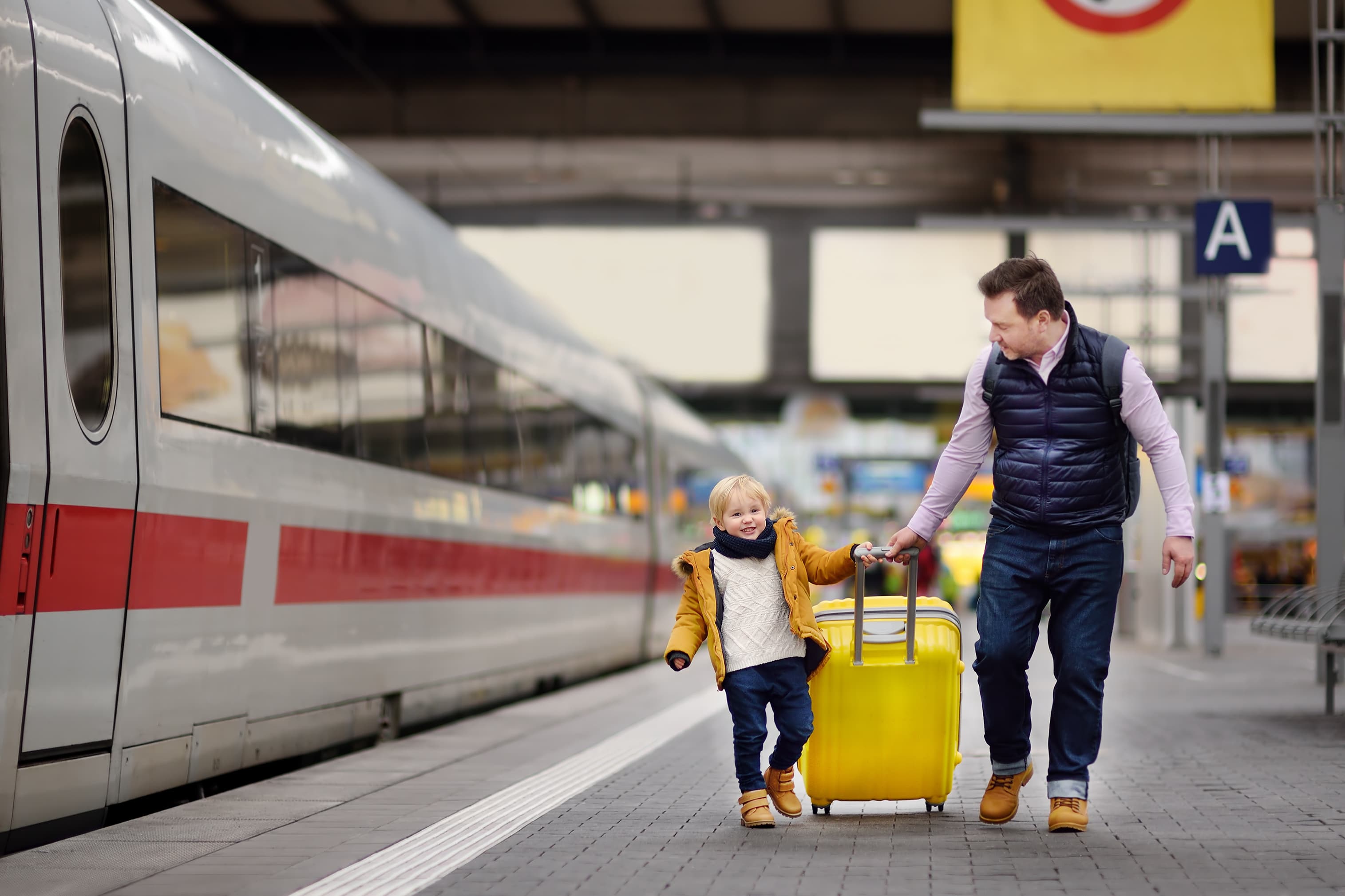 A man and a child with a yellow suitcase walk along a train platform beside a stationary train.