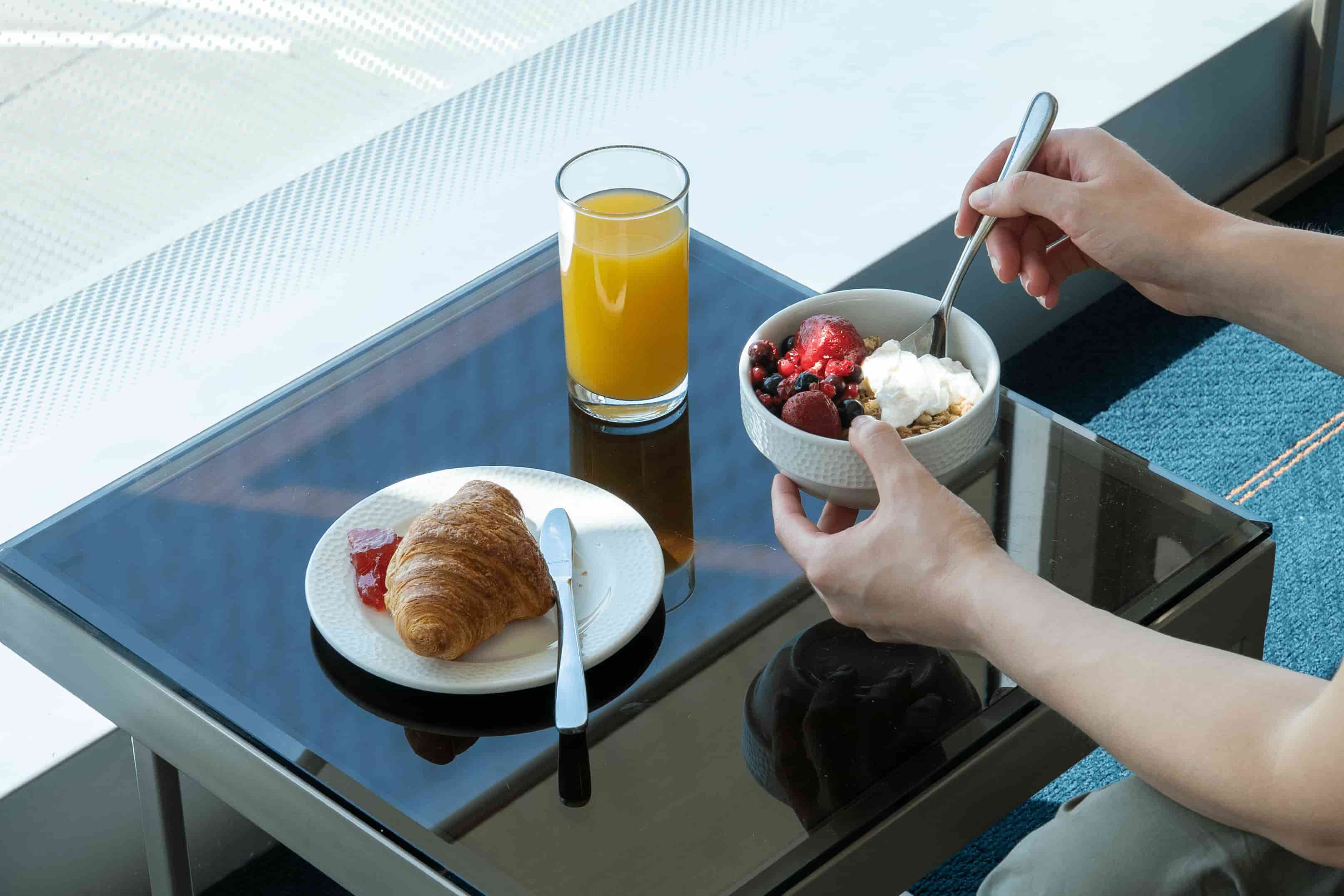 A person holds a bowl of granola with yogurt and berries beside a glass of orange juice and a plate with a croissant and jam on a glass table.