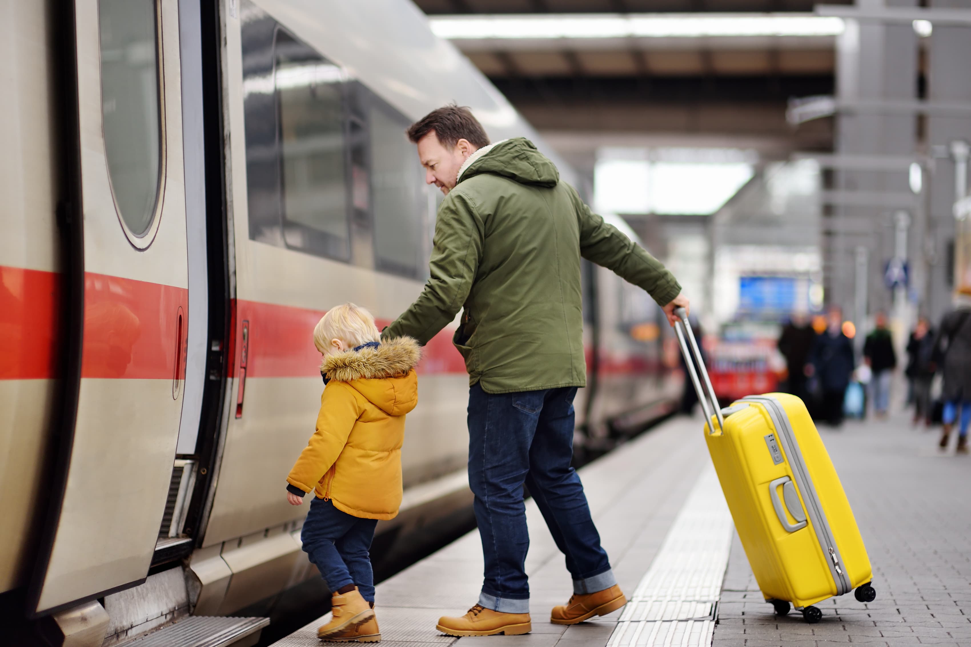 Man and child boarding a train at a station; man holds a yellow suitcase and the child's hand, both in winter jackets.