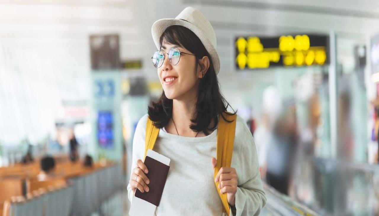 Woman with a hat and glasses holding a passport and boarding pass at an airport, with a yellow backpack and blurred sign in the background.