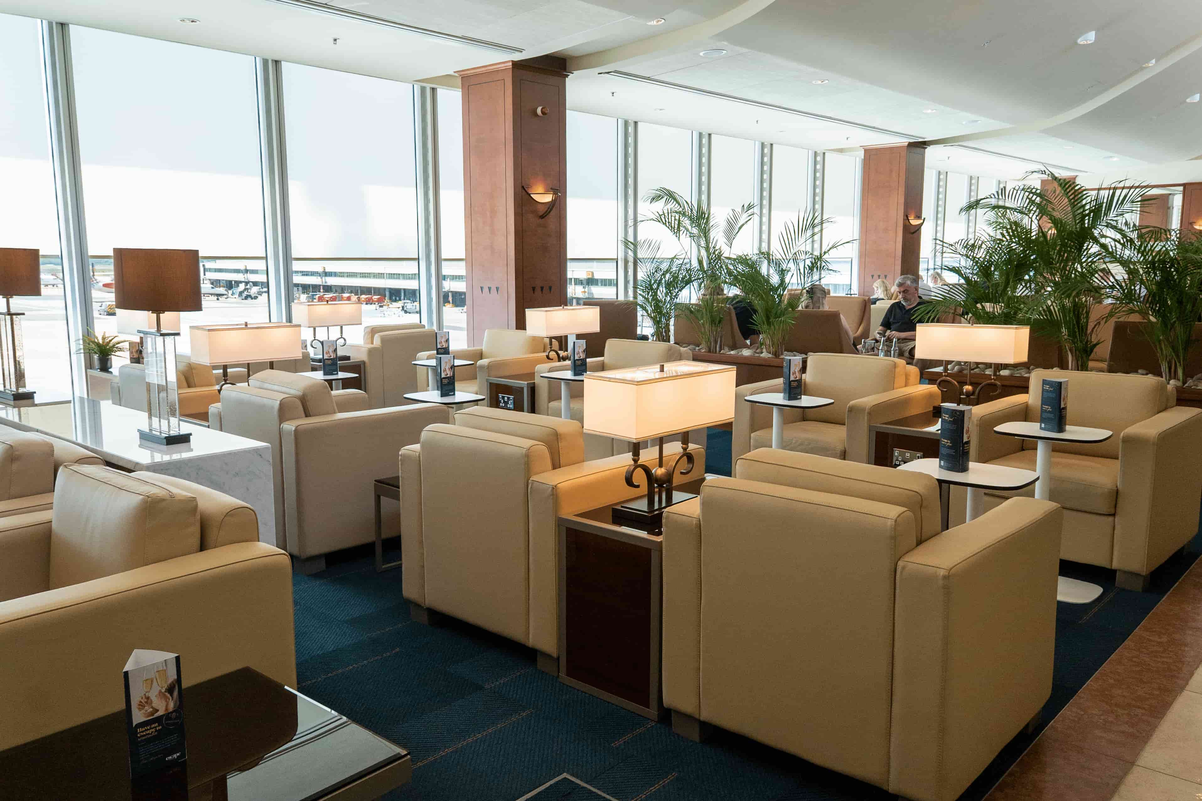 Luxurious airport lounge with beige armchairs, small tables, and elegant lamps. Large windows offer views of the runway and parked aircraft.