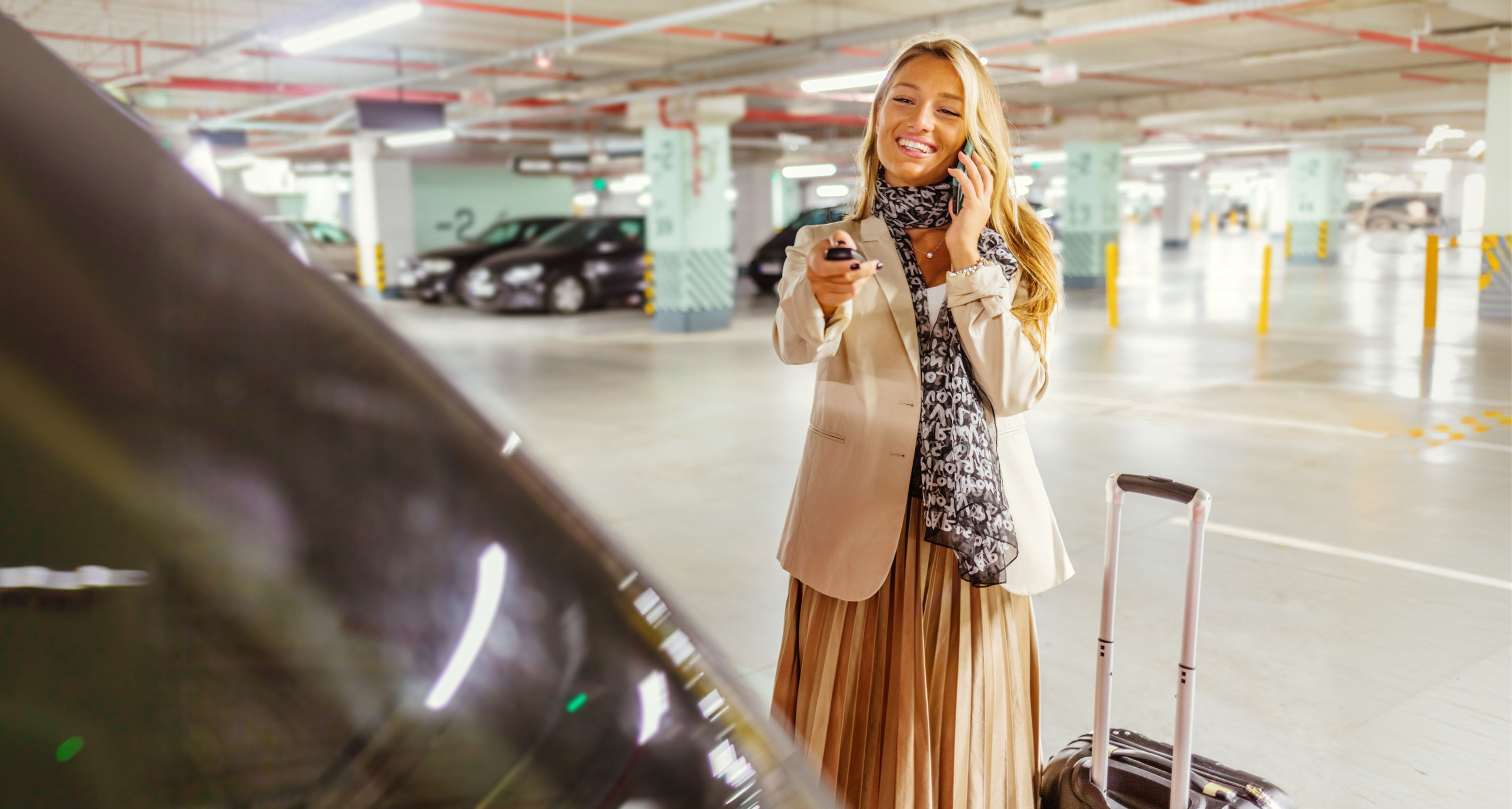 Woman in a parking garage smiling, talking on a phone, holding car keys, with luggage by her side.