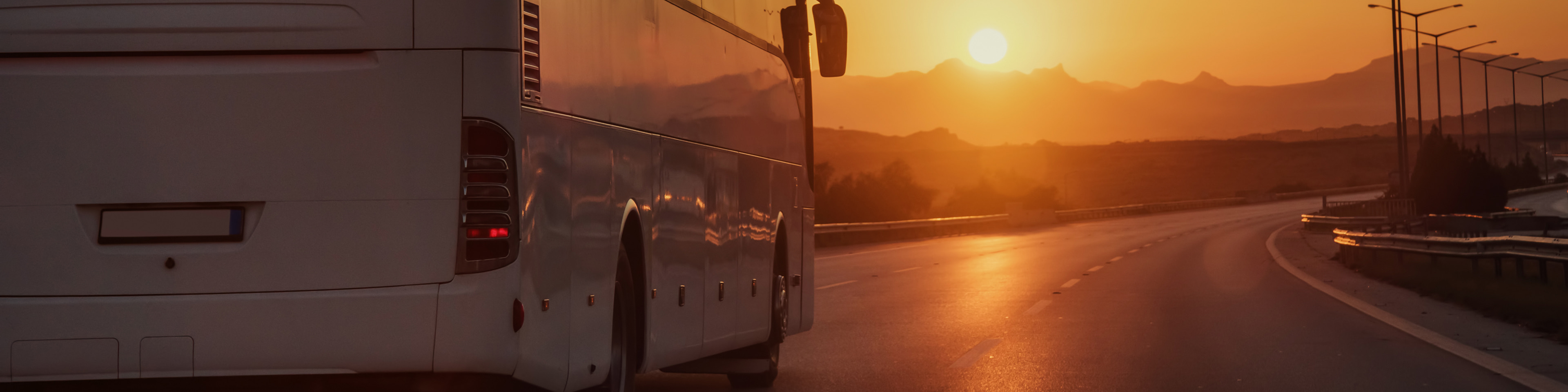A bus travels on an empty highway during a sunset, with orange and pink hues illuminating the sky and distant mountains.