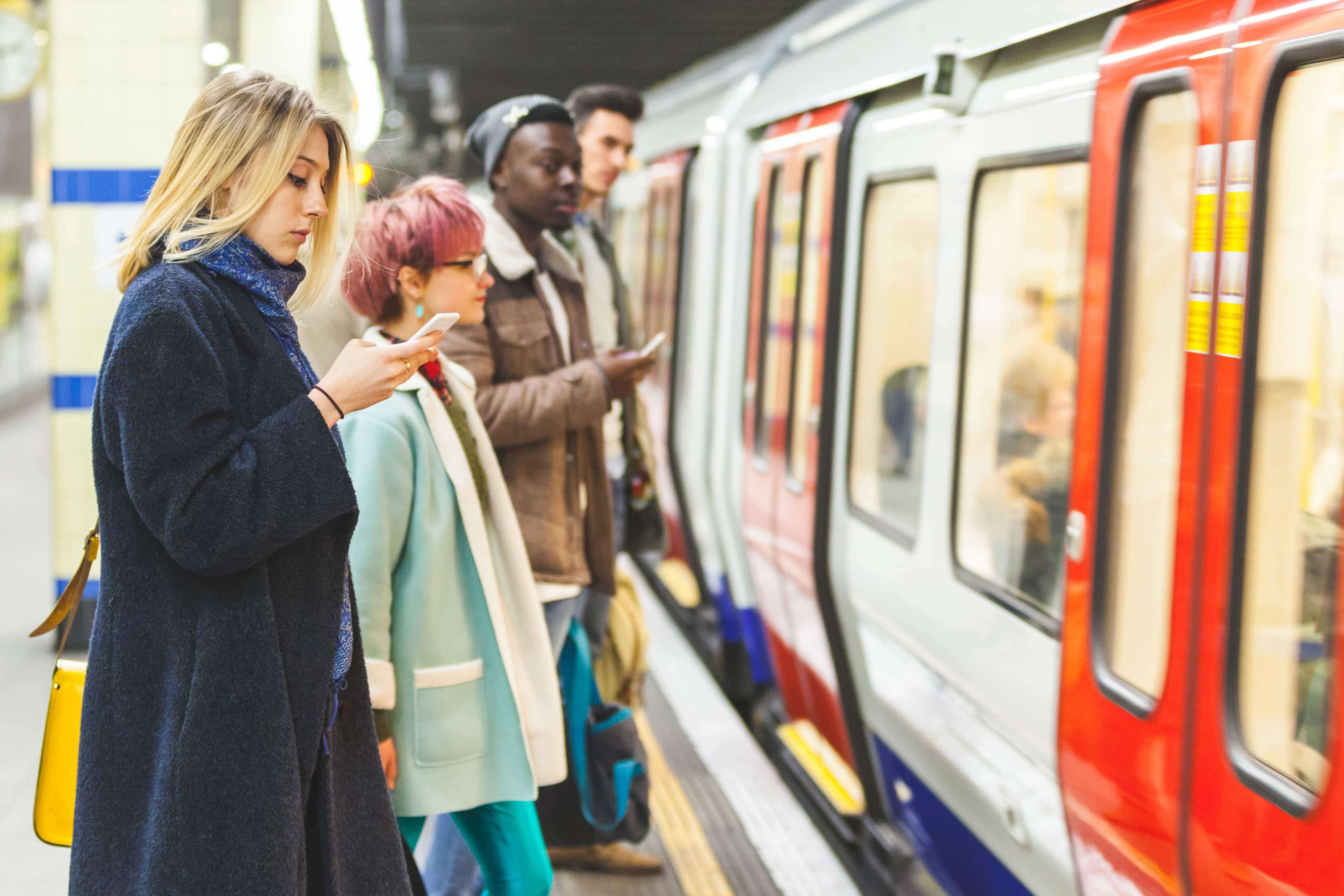 People standing on a subway platform, looking at their phones, as a train arrives.