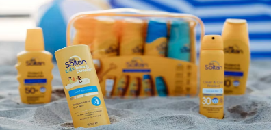 Assorted Soltan sunscreen products on sand with a beach background, including bottles and a clear bag set containing multiple sunscreens.