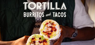 Person holding a halved burrito filled with ingredients, under a sign reading "Tortilla Burritos and Tacos."