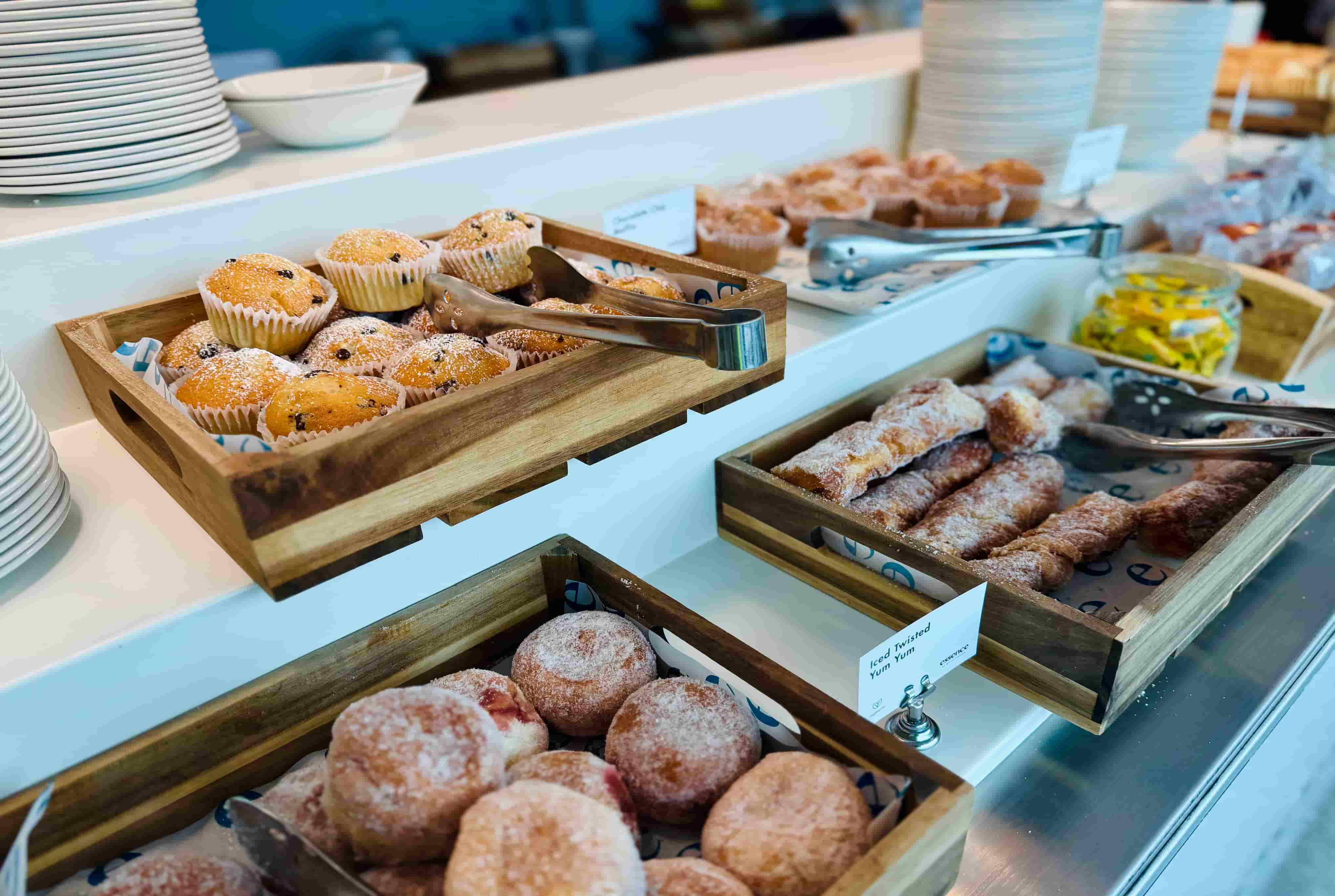 Assorted pastries, including muffins and donuts, displayed in wooden trays, with tongs and stacked plates in the background.
