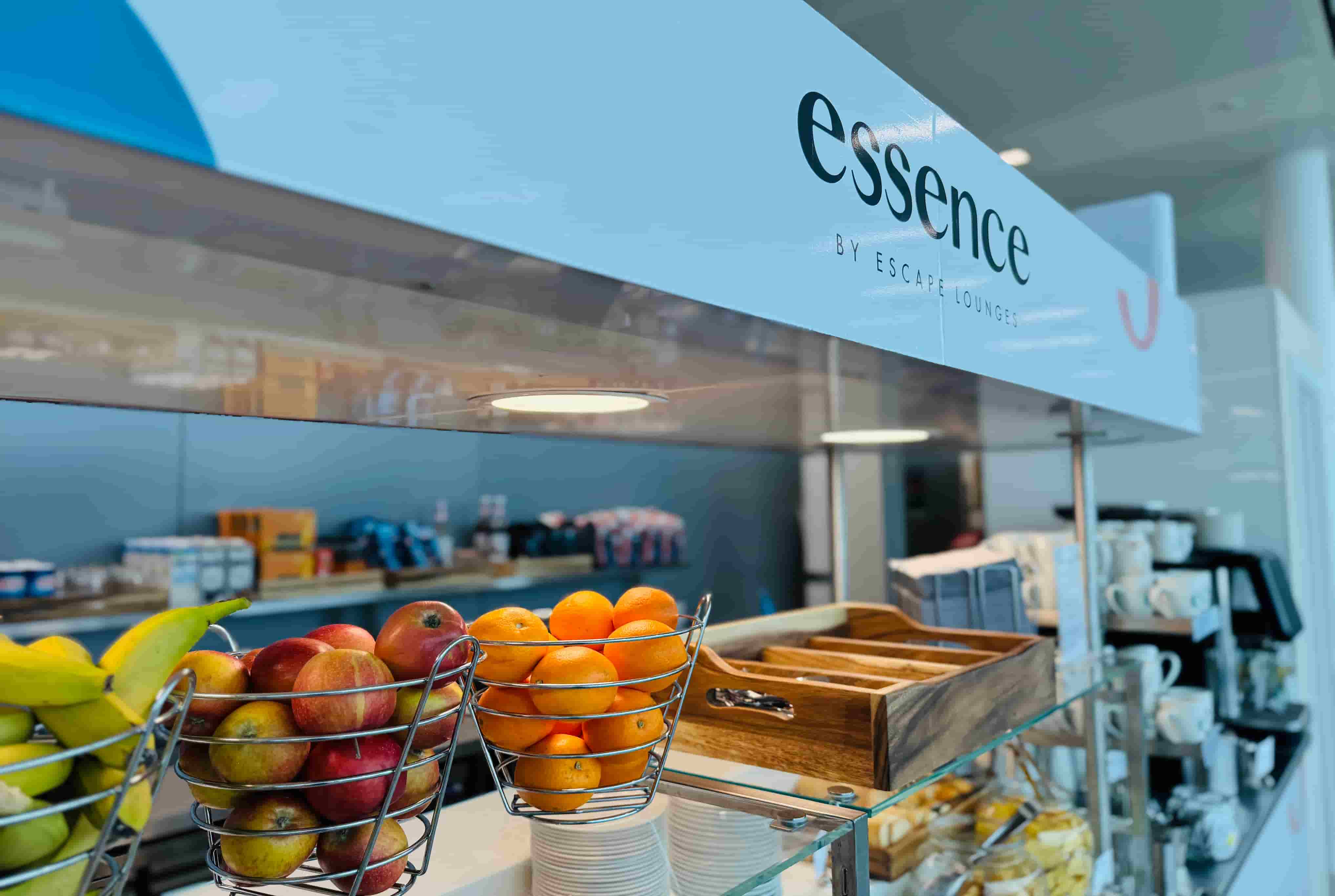 Fruits in wire baskets, cups, and cutlery on a buffet at a lounge labeled "essence by Escape Lounges."