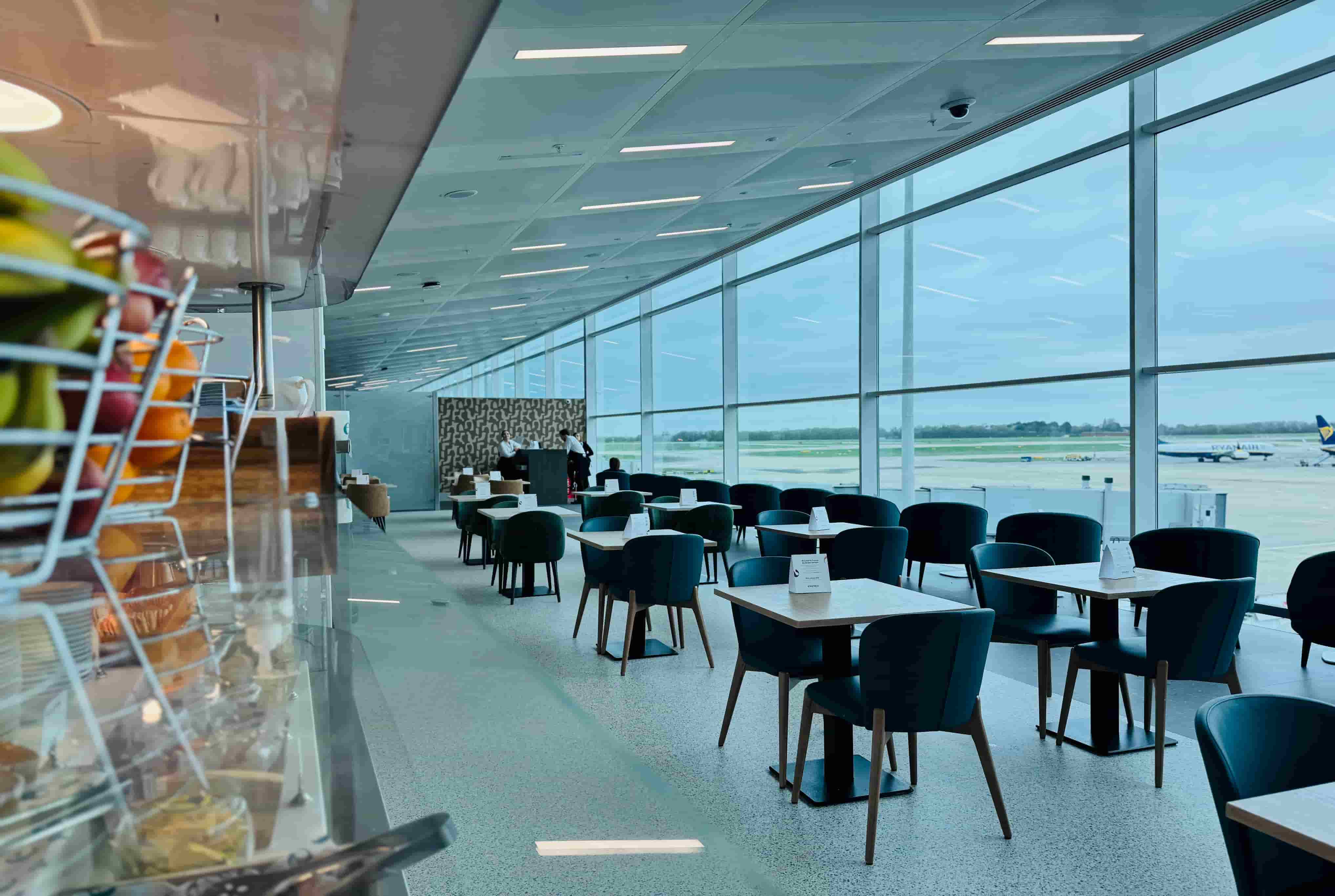 Airport lounge with empty tables and chairs, large windows showing airplanes on the runway, and a fruit display in the foreground.