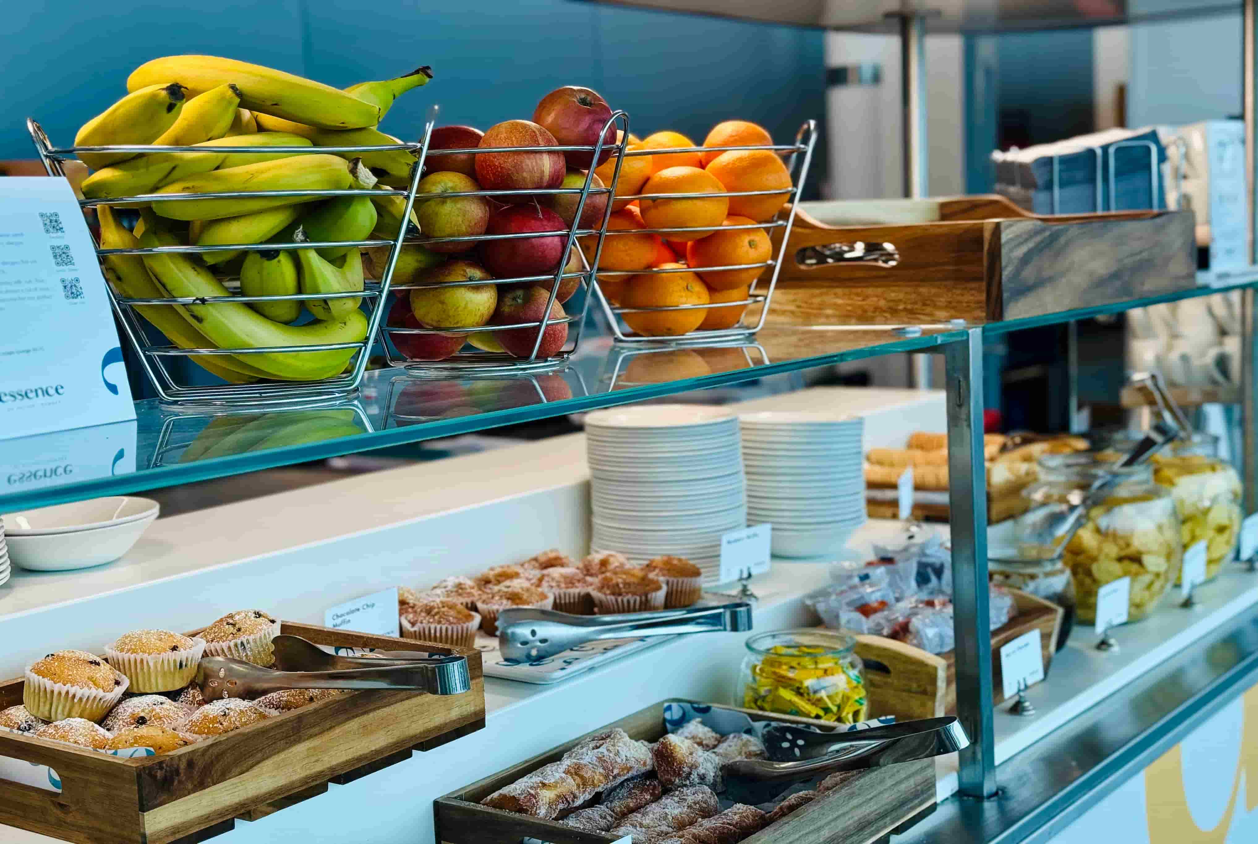 A cafe counter displaying baskets of bananas, apples, oranges, muffins, pastries, and snacks with plates stacked in the background.