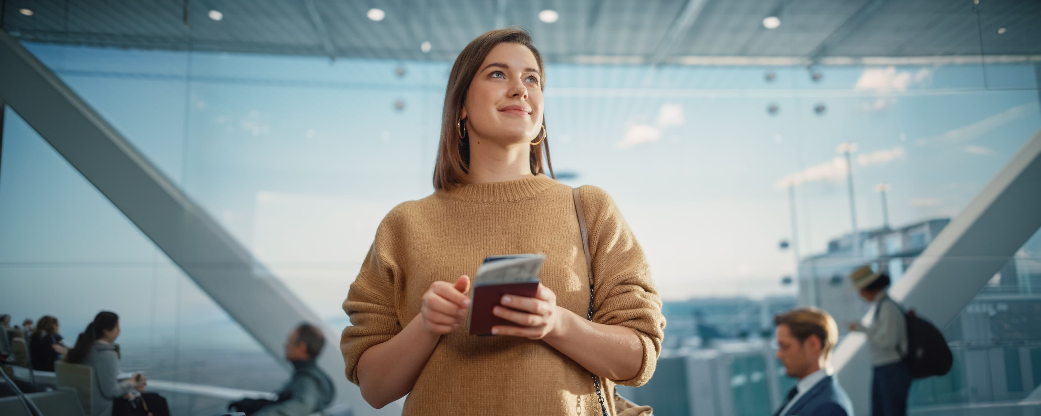 A woman in a brown jumper holding a passport and phone stands in a modern airport, with blurred travellers in the background.